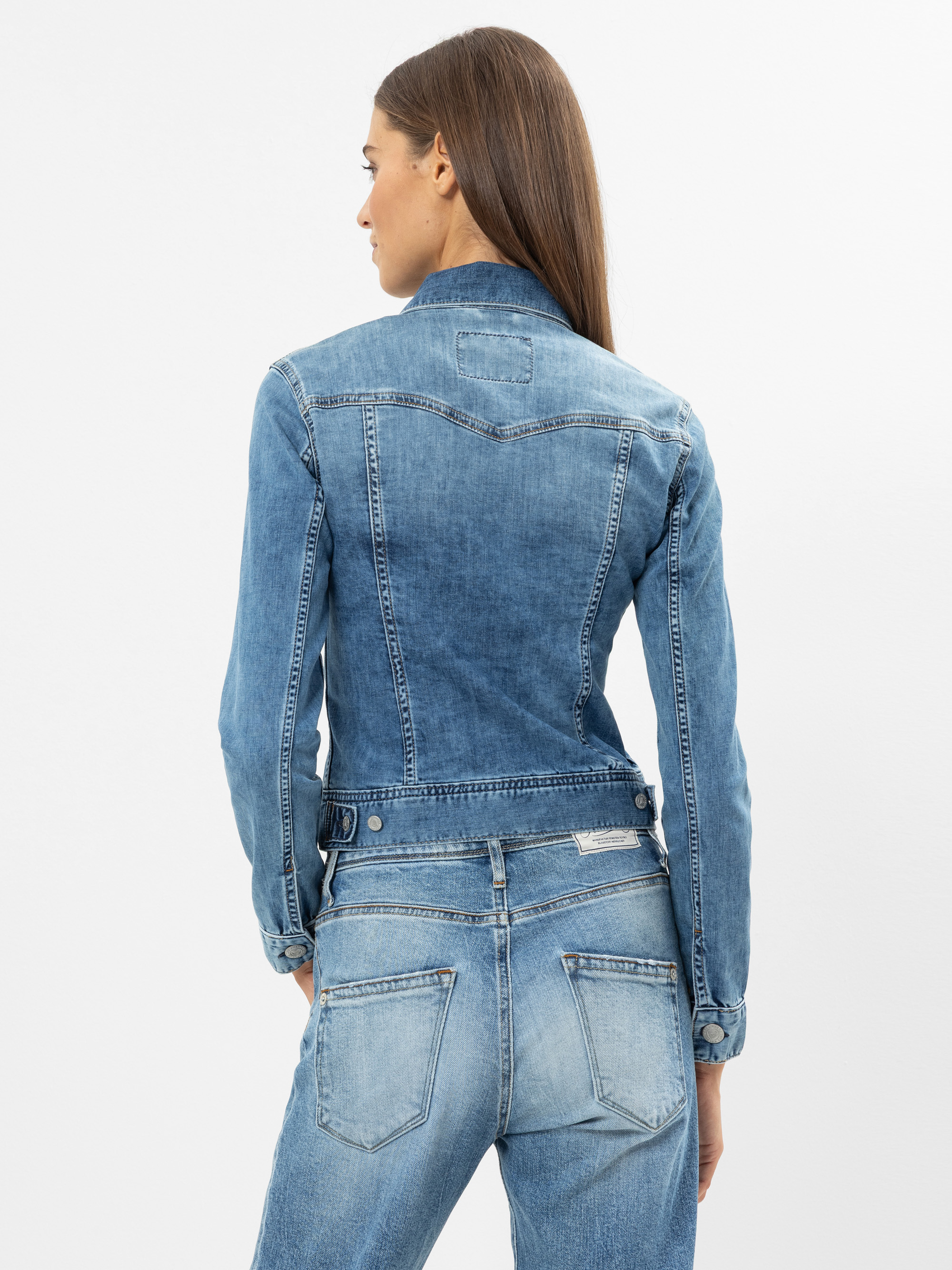 Man sieht eine Frau mit glattem braunem Haar von hinten, die eine hellblaue Jeansjacke und eine dazu passende Jeans vor einem schlichten weißen Hintergrund trägt.