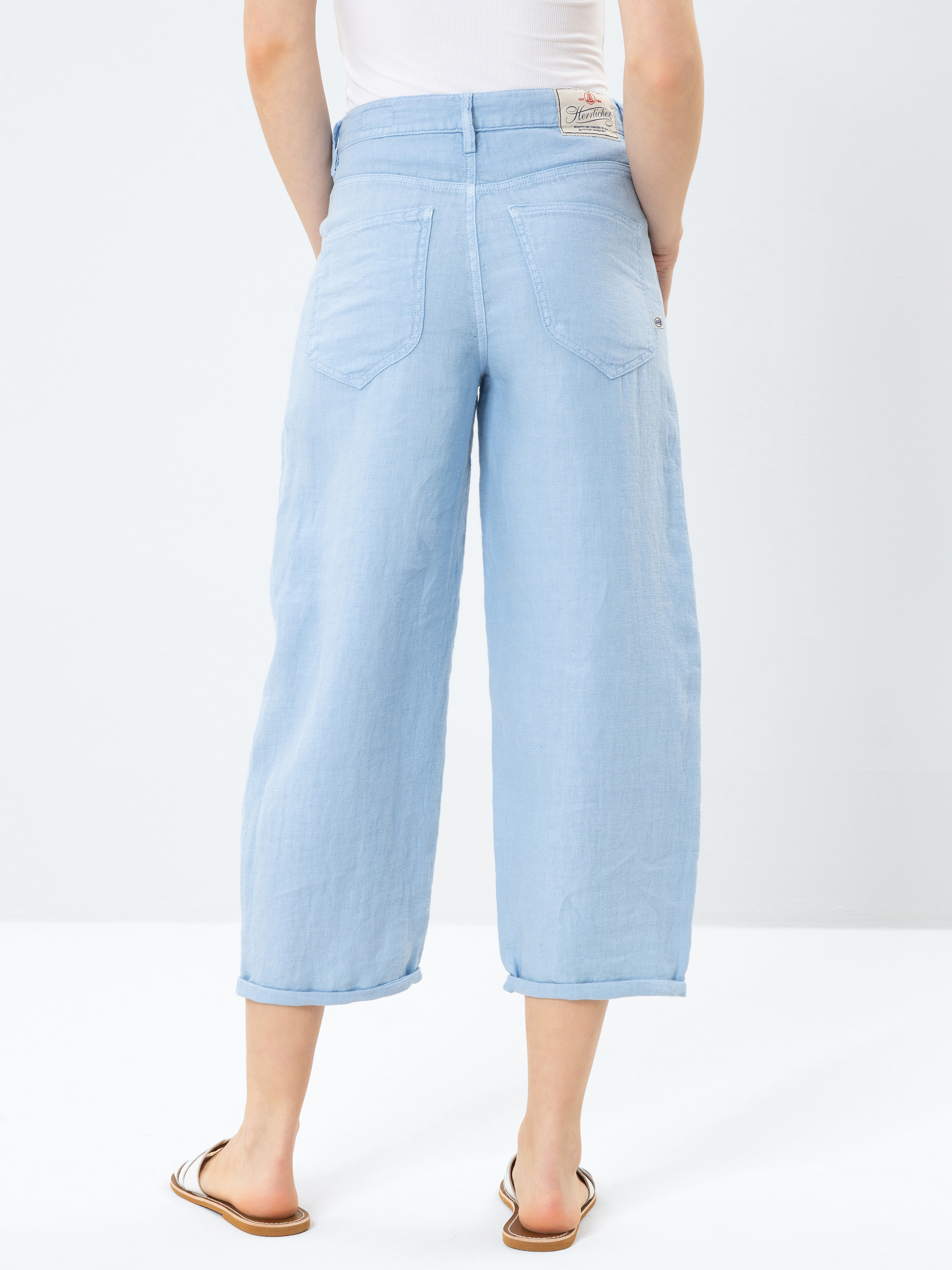 Stehende Person vor weißem Hintergrund, die hellblaue, weit geschnittene Jeans, ein weißes, ärmelloses Oberteil und braune Sandalen trägt. Das Bild ist von hinten aufgenommen und zeigt die hinteren Details der Jeans.