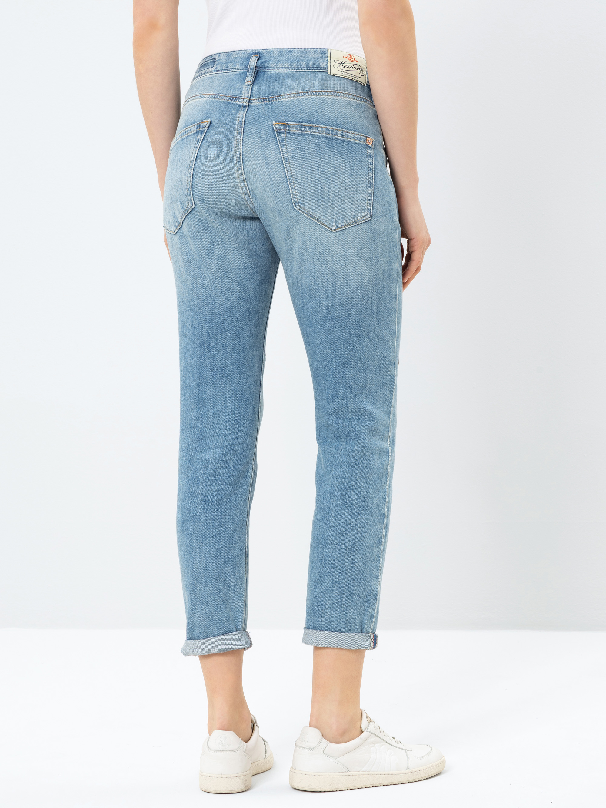 Person in hellblauen Jeans mit Bündchen und weißen Turnschuhen, die mit dem Rücken zur Kamera vor einem schlichten hellen Hintergrund steht.