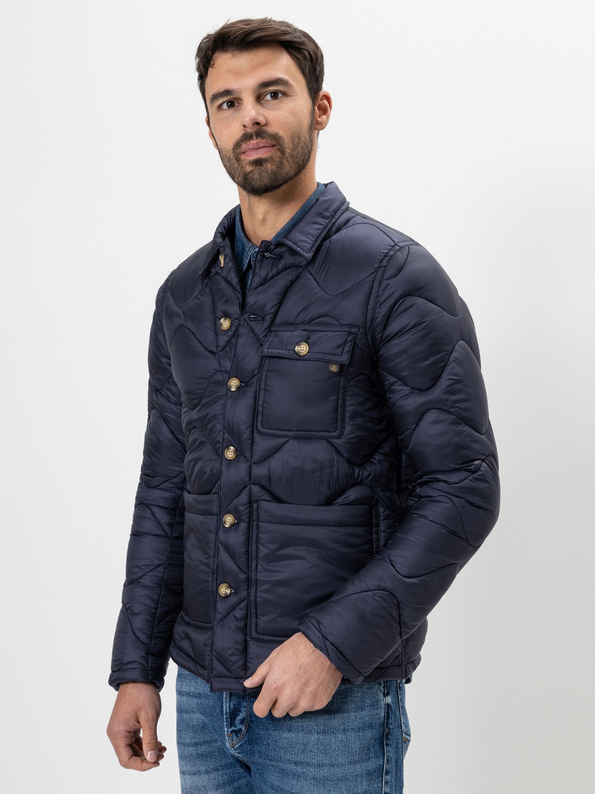Ein Mann mit Bart, der eine dunkelblaue Steppjacke mit Knöpfen und blaue Jeans trägt, steht vor einem schlichten weißen Hintergrund und schaut leicht zur Seite.