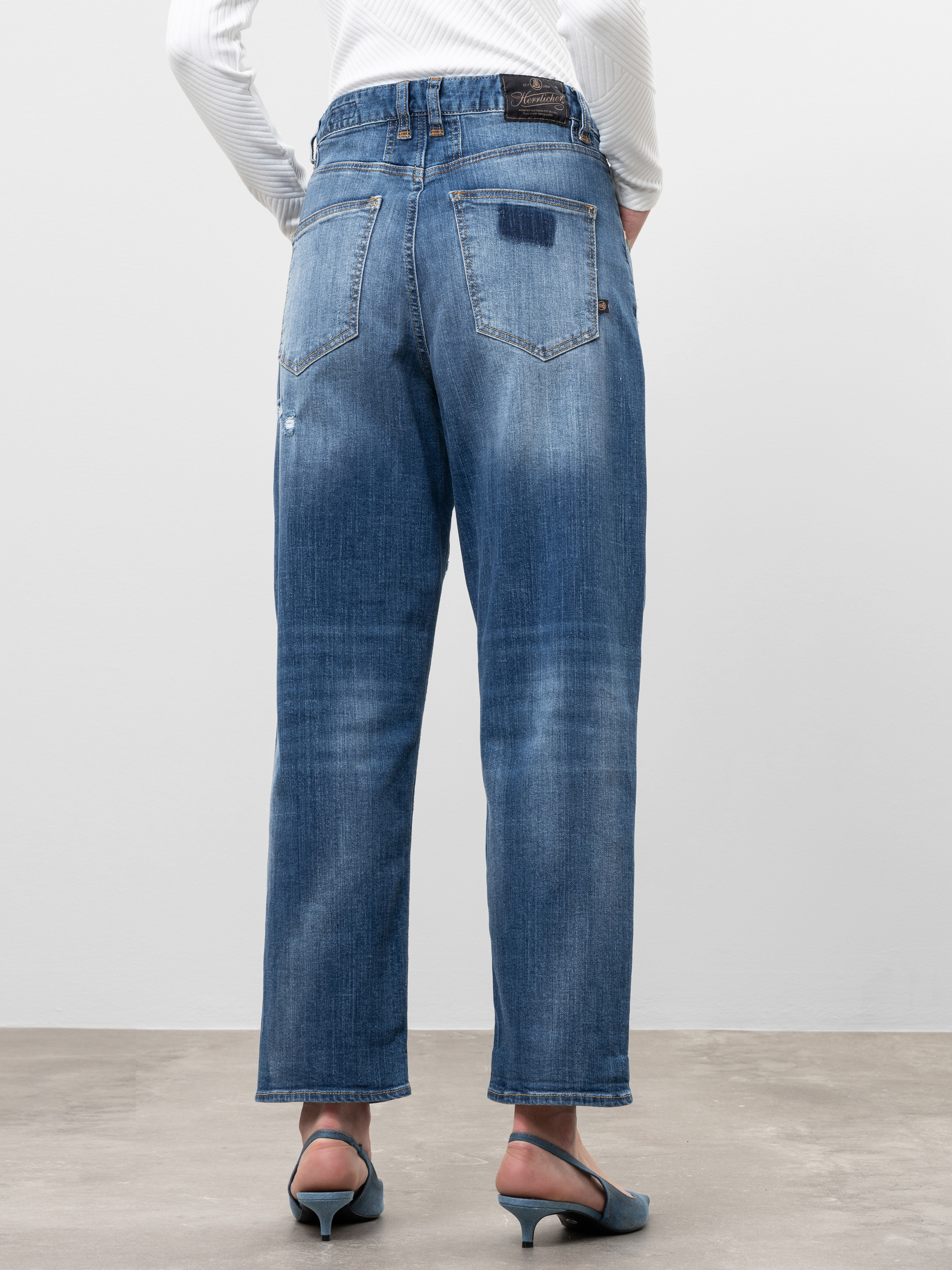 Eine Person, die blaue Jeans mit geradem Bein und blaugrüne Sandalen mit Absatz trägt, steht mit dem Rücken zur Kamera vor einem schlichten Hintergrund. Die Jeans hat verblasste Details und zwei Gesäßtaschen.