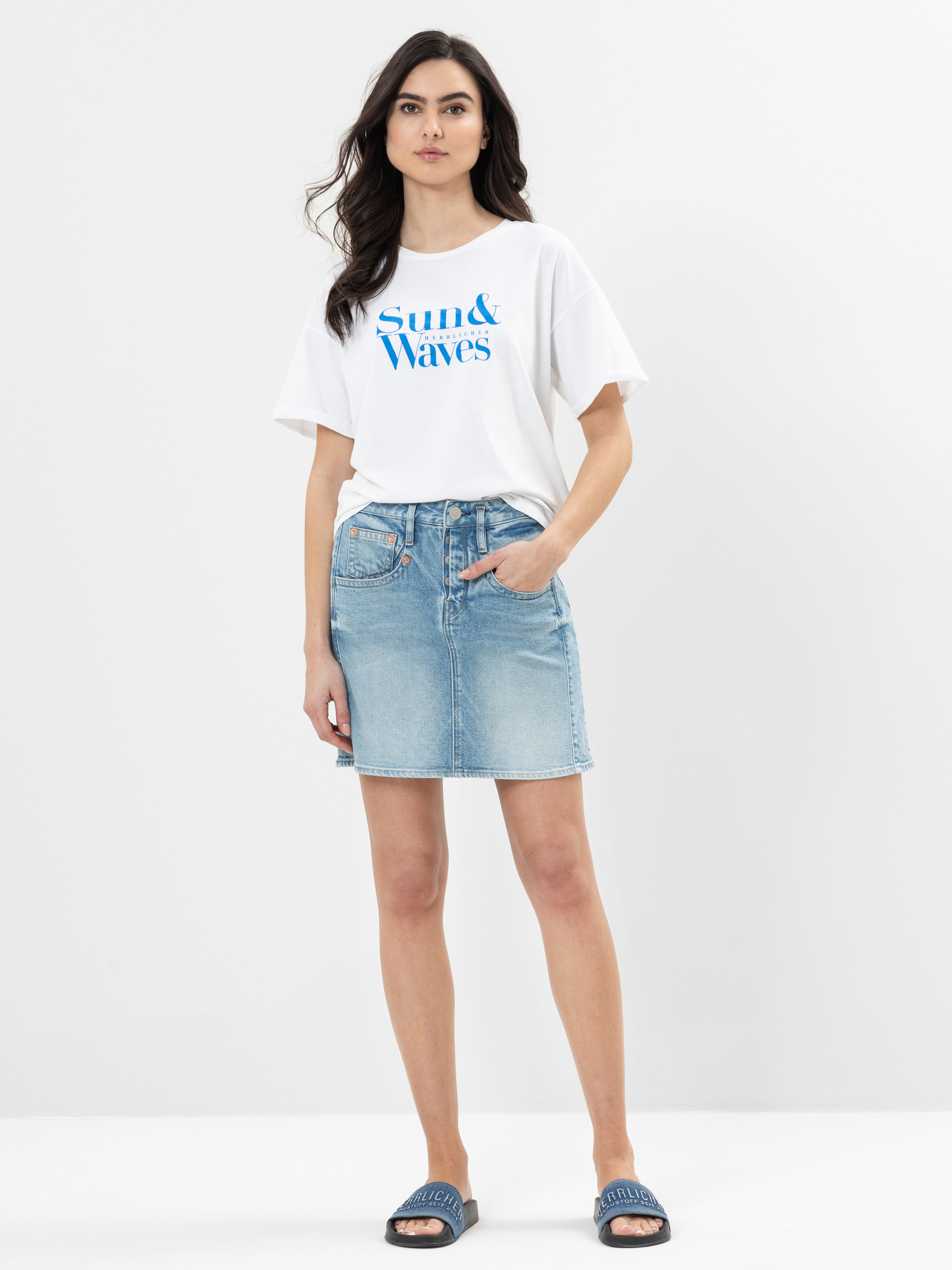 Eine Frau steht vor einem weißen Hintergrund und trägt ein weißes Sun & Waves-T-Shirt, einen hellblauen Jeansrock und blaue Slide-Sandalen. Sie hat lange dunkle Haare und eine Hand in der Tasche.