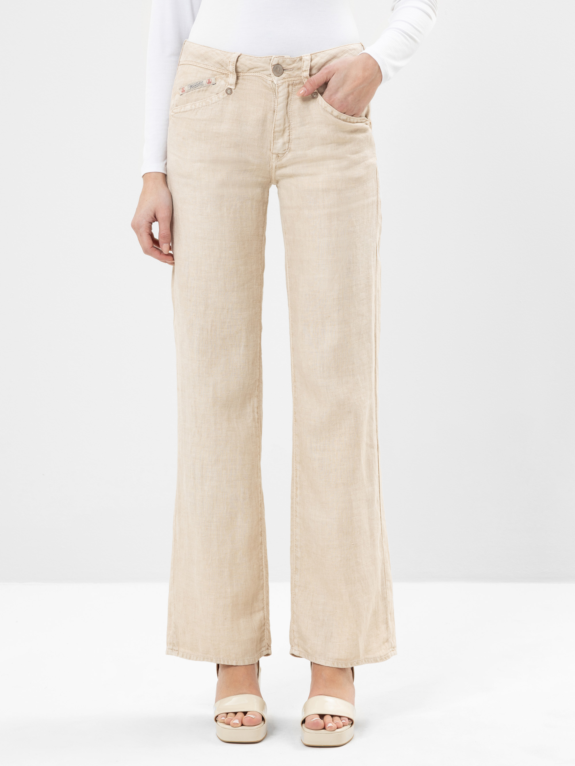 Eine Person, die eine hellbeige Jeans mit weitem Bein und ein weißes, langärmeliges Oberteil trägt, eine Hand in einer Tasche hat und beigefarbene, offene Sandalen trägt.