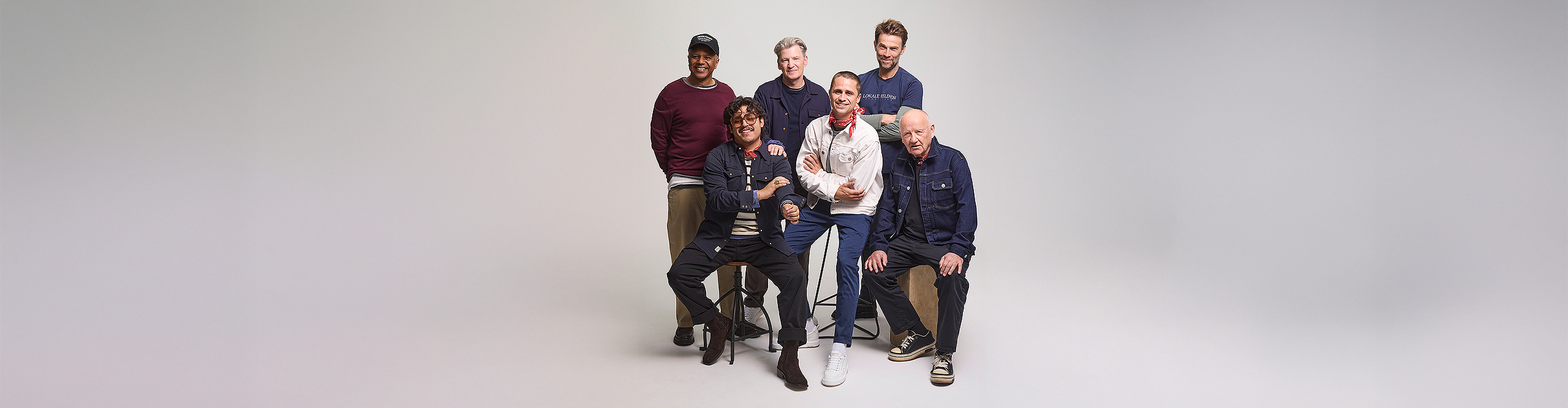 Sechs Männer posieren gemeinsam in einem Studio vor einem schlichten hellgrauen Hintergrund; vier stehen, zwei sitzen vorne, sie sind leger gekleidet und lächeln.