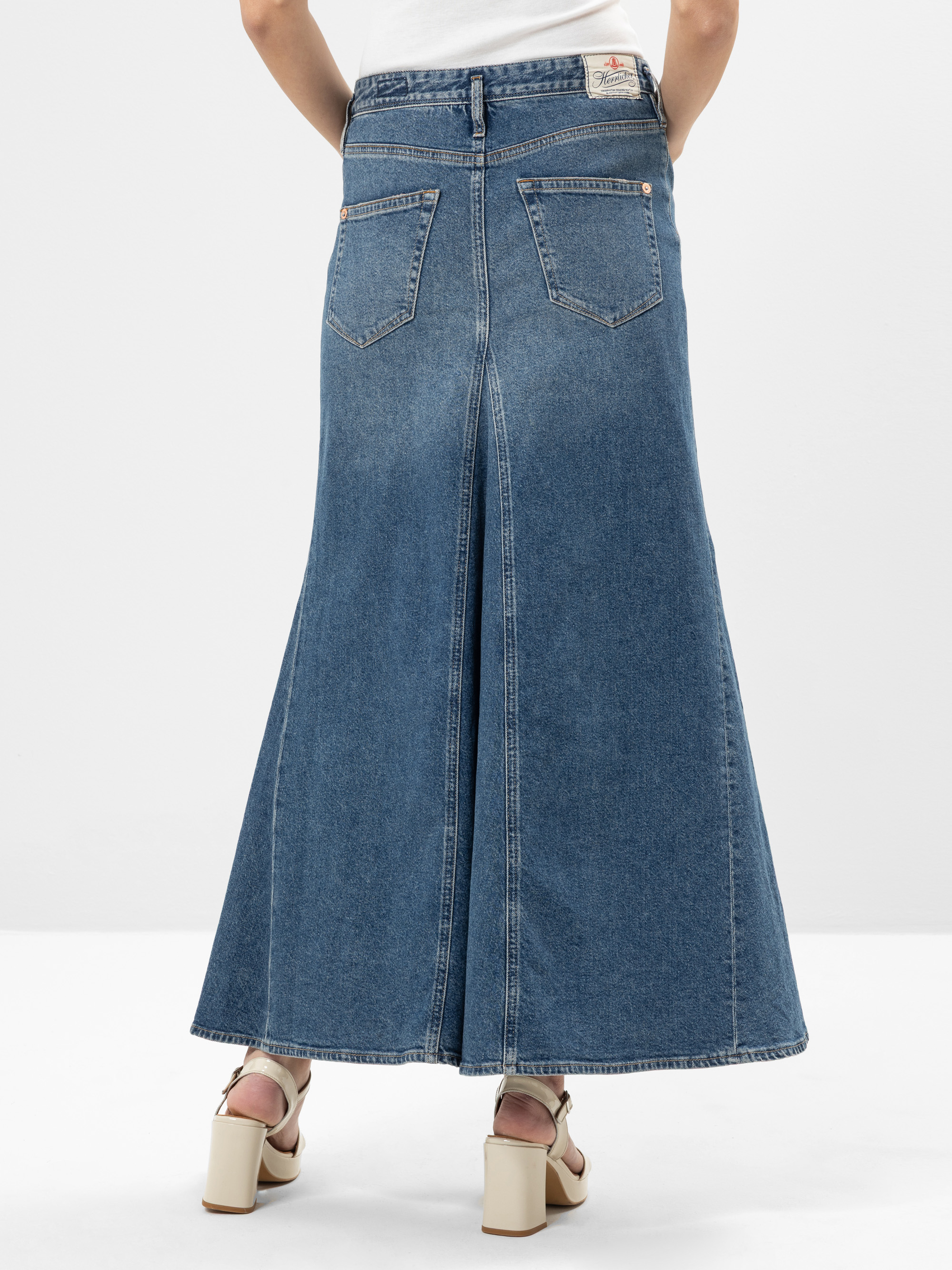 Eine Person, die einen langen, ausgestellten blauen Jeansrock mit Gesäßtaschen und weißen hochhackigen Sandalen trägt, wird von hinten vor einem schlichten weißen Hintergrund gezeigt.