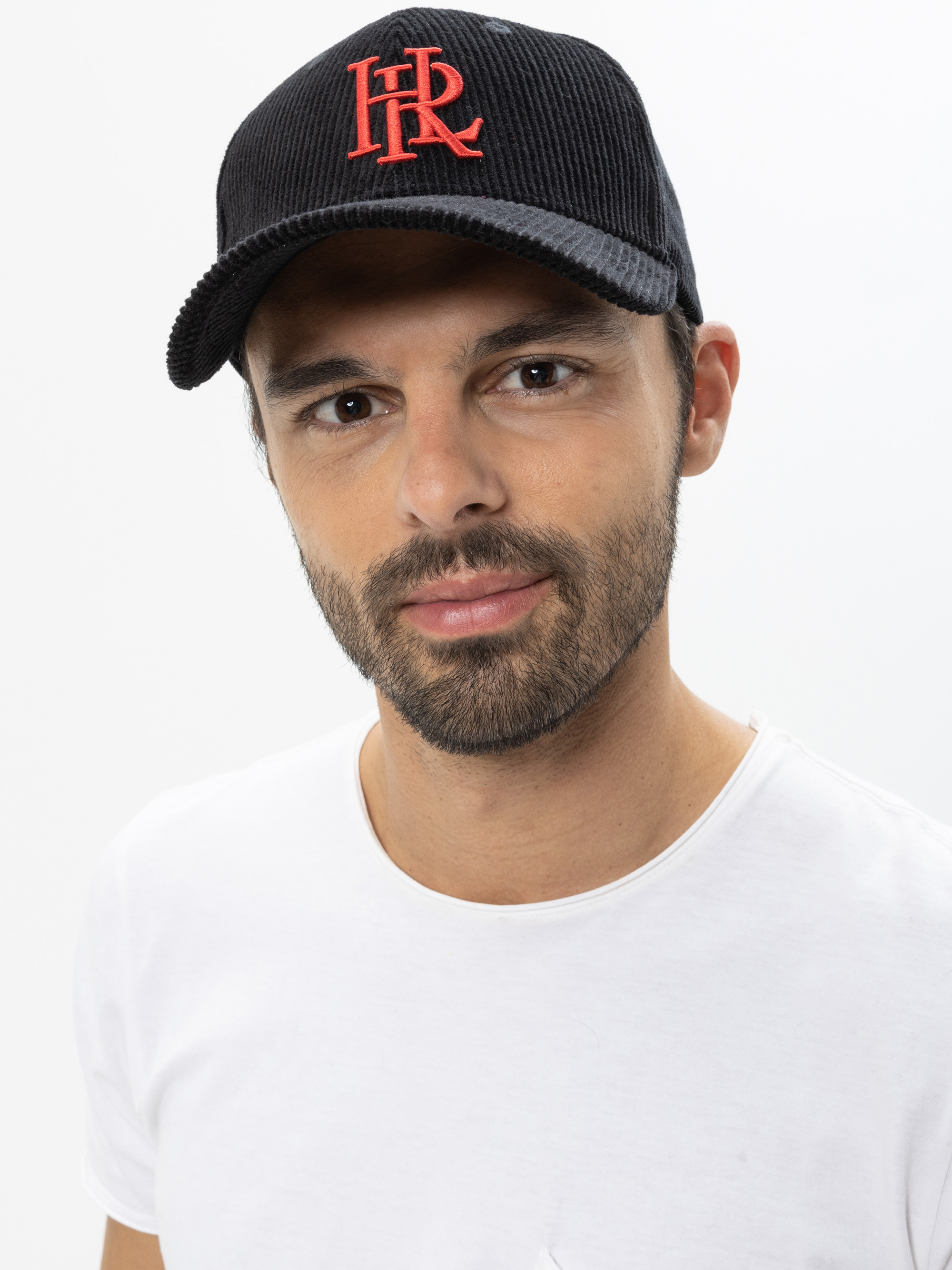 Ein Mann mit kurzem Bart und Schnurrbart, der eine schwarze Kappe mit roten HR-Initialen und ein weißes T-Shirt trägt, steht vor einem schlichten weißen Hintergrund.