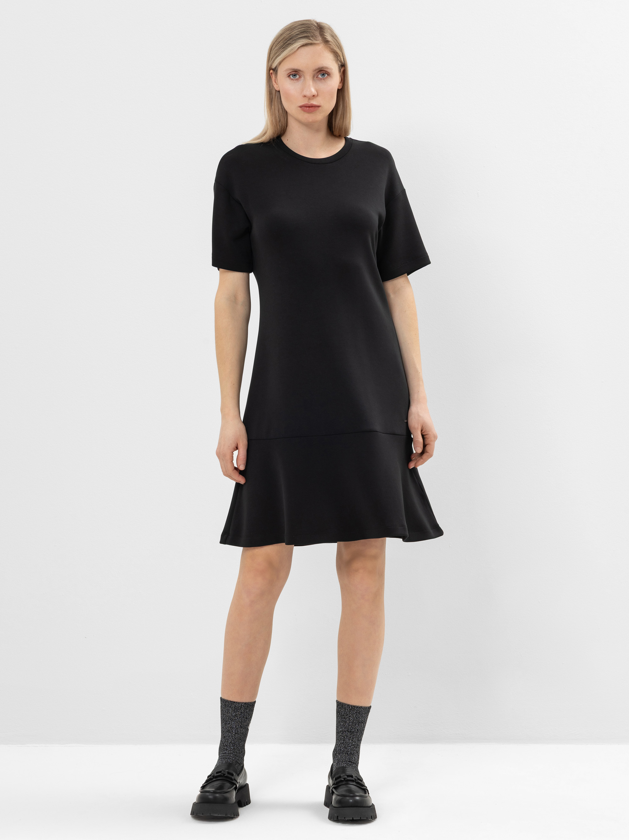 Eine Frau steht vor einem schlichten weißen Hintergrund und trägt ein kurzärmeliges, schwarzes, locker sitzendes Kleid mit tiefer Taille und ausgestelltem Saum. Sie kombiniert das Kleid mit grauen Socken und schwarzen Schuhen.