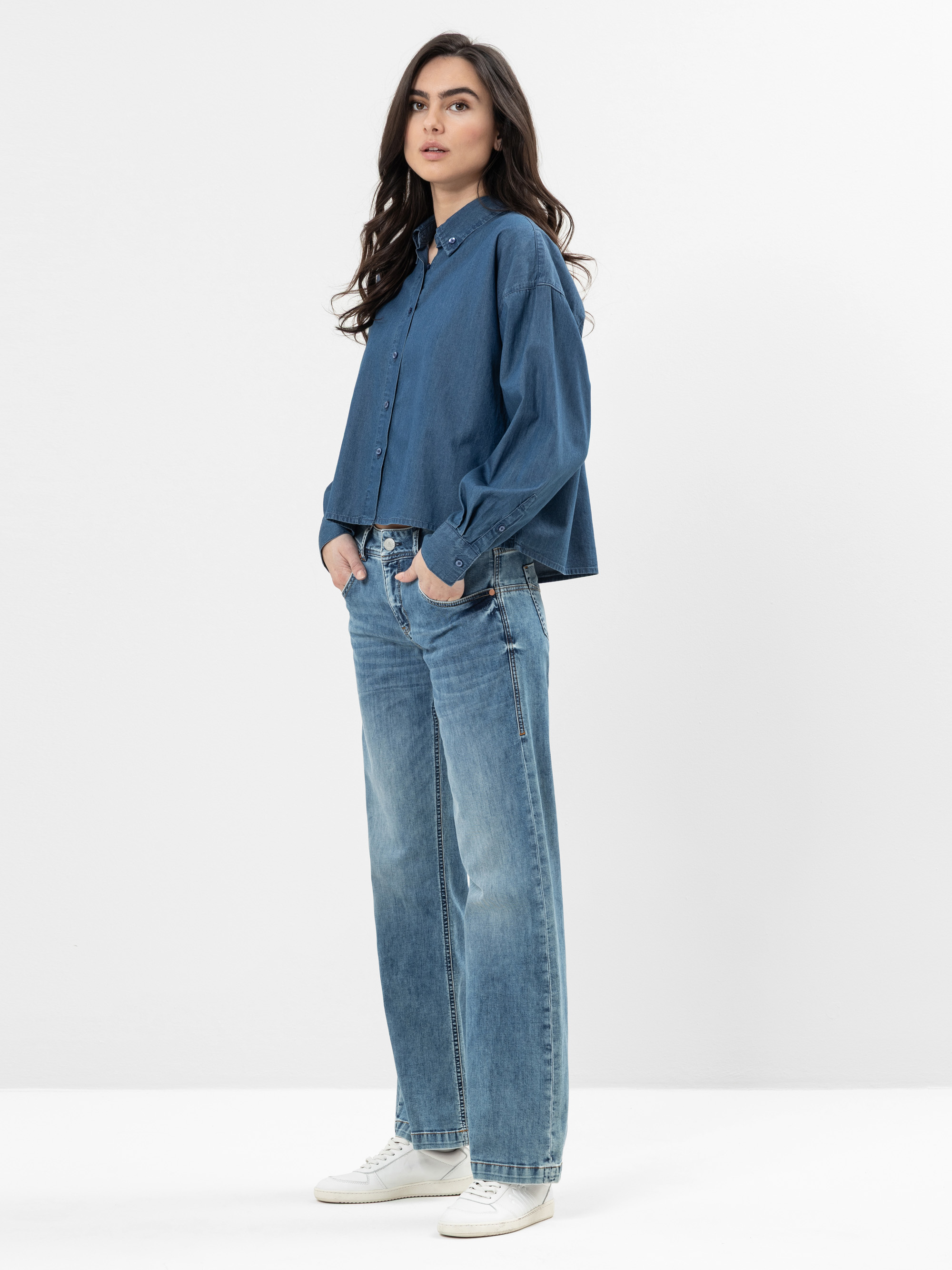 Eine Frau mit langen dunklen Haaren steht vor einem schlichten weißen Hintergrund. Sie trägt ein lockeres blaues Button-up-Hemd, eine weit geschnittene Bluejeans und weiße Turnschuhe. Sie hat die Hände in den Taschen und blickt leicht zur Seite.