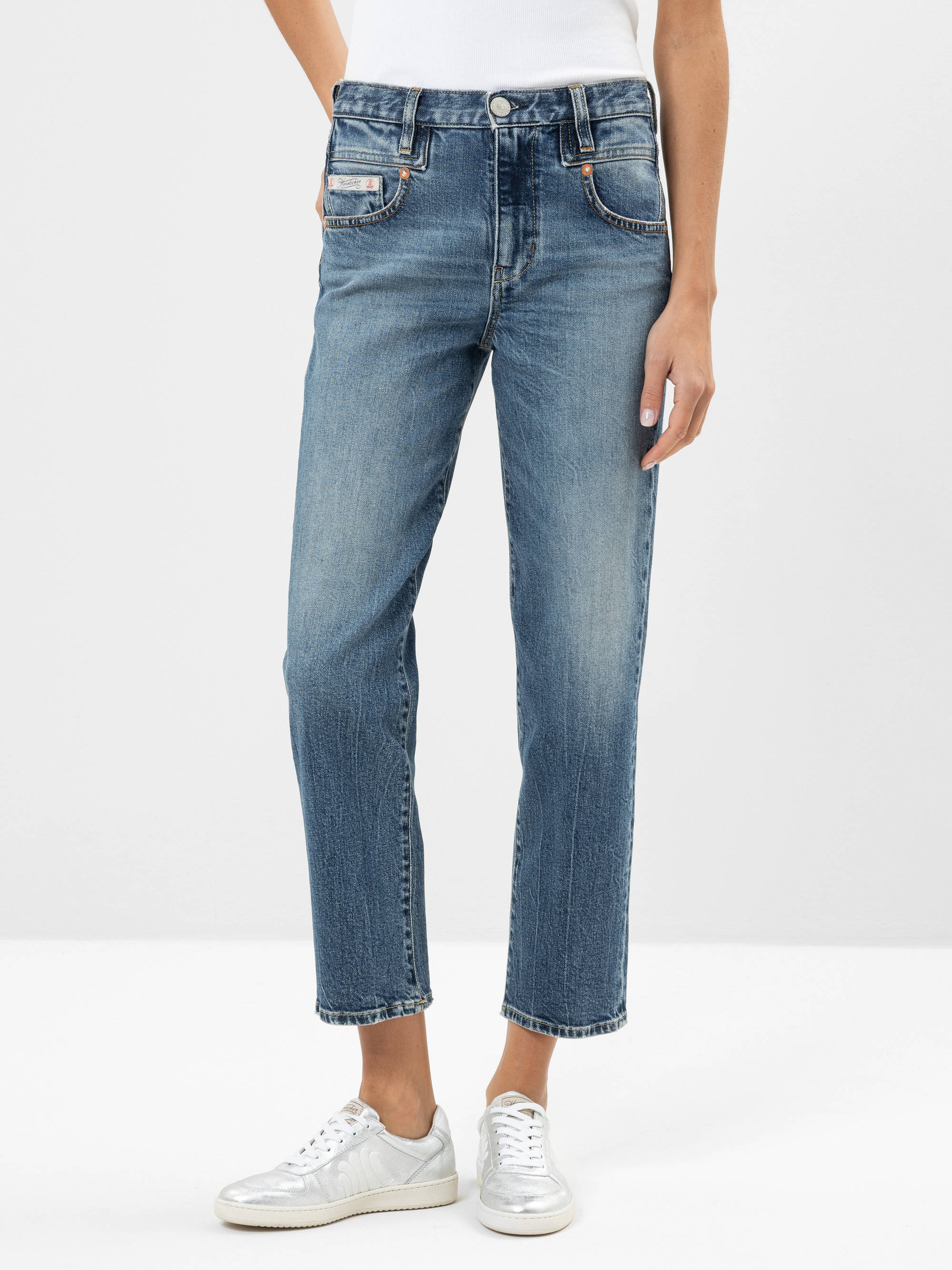 Eine Person, die blaue Jeans mit geradem Bein, ein weißes T-Shirt und silberne Turnschuhe trägt, steht vor einem schlichten, hellen Hintergrund. Nur der Körper von den Schultern abwärts ist sichtbar.