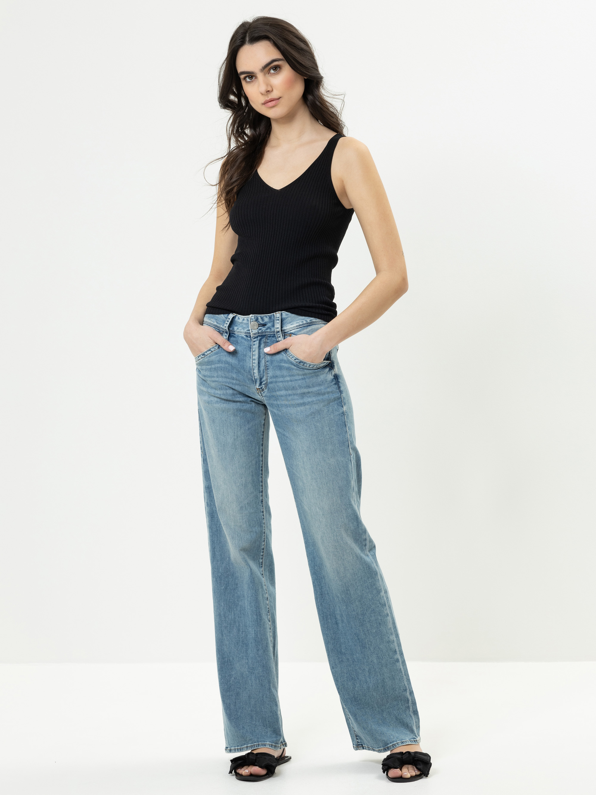 Frau, die vor einem weißen Hintergrund steht, ein ärmelloses schwarzes geripptes Oberteil, hellblaue Jeans mit weitem Bein und schwarze Sandalen trägt, die Hände in den Taschen und einen neutralen Gesichtsausdruck hat.