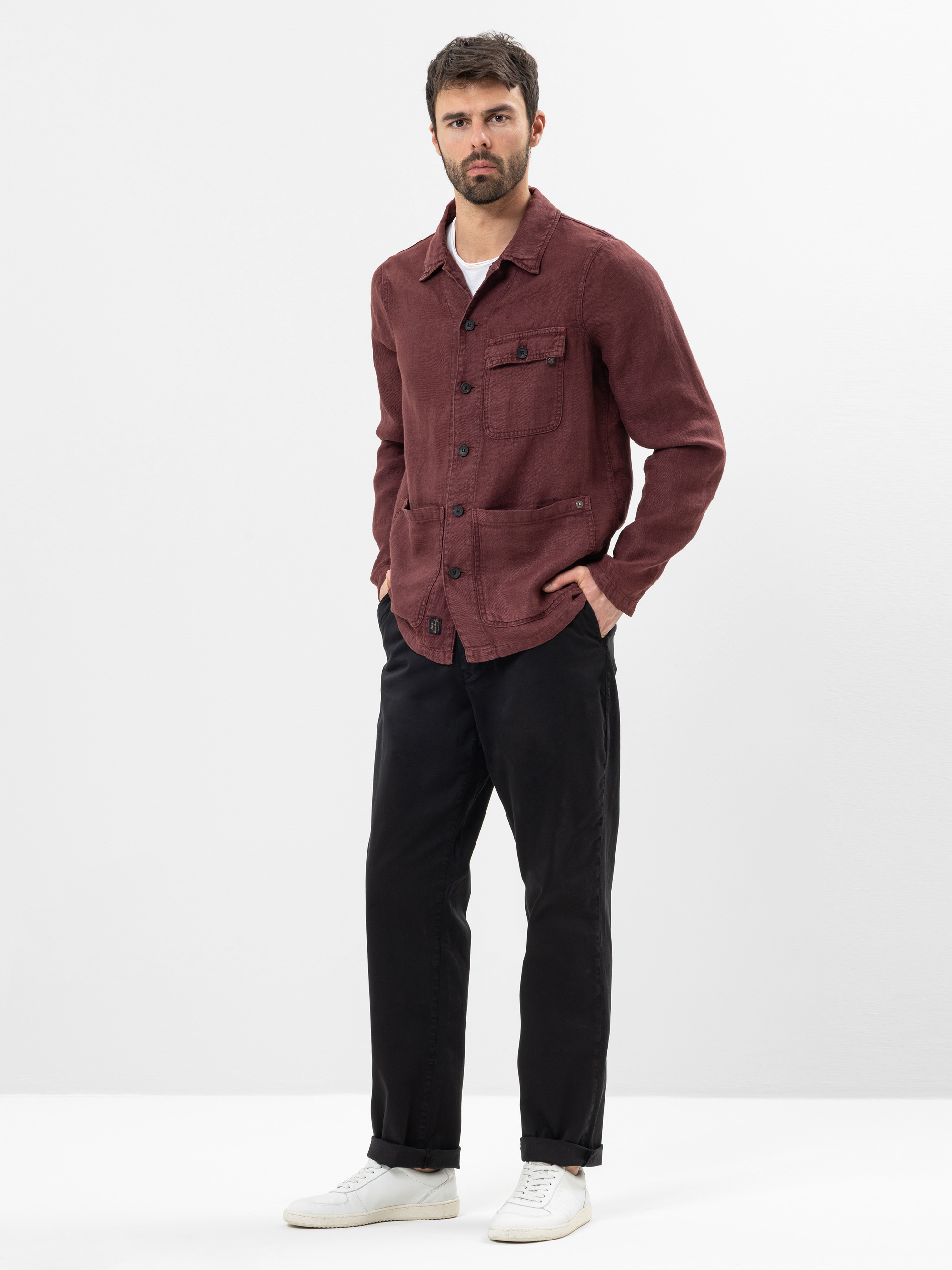 Ein Mann mit kurzen dunklen Haaren und einem Bart steht vor einem schlichten weißen Hintergrund. Er trägt eine kastanienbraune Button-up-Jacke, eine schwarze Hose, ein weißes Hemd und weiße Turnschuhe und hat die Hände in den Taschen.