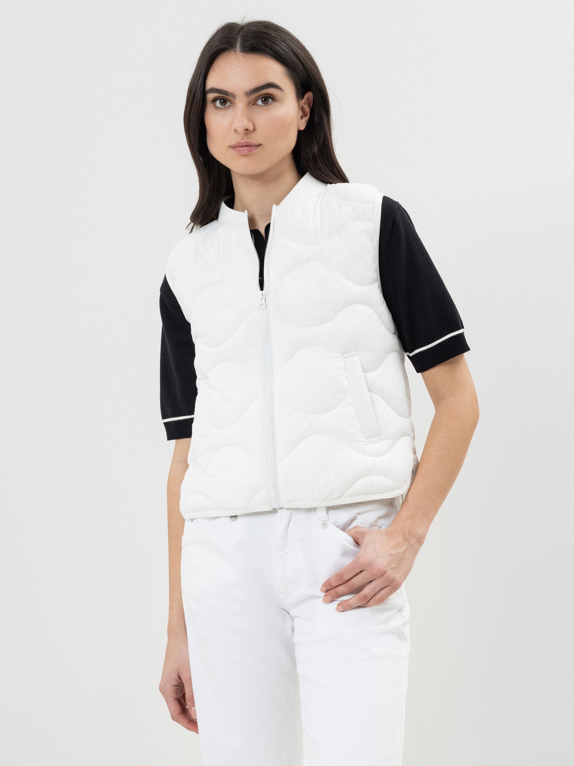 Eine Frau mit glattem dunklem Haar trägt eine weiße Steppweste über einem kurzärmeligen schwarzen Polohemd und einer weißen Hose und steht vor einem schlichten hellen Hintergrund.