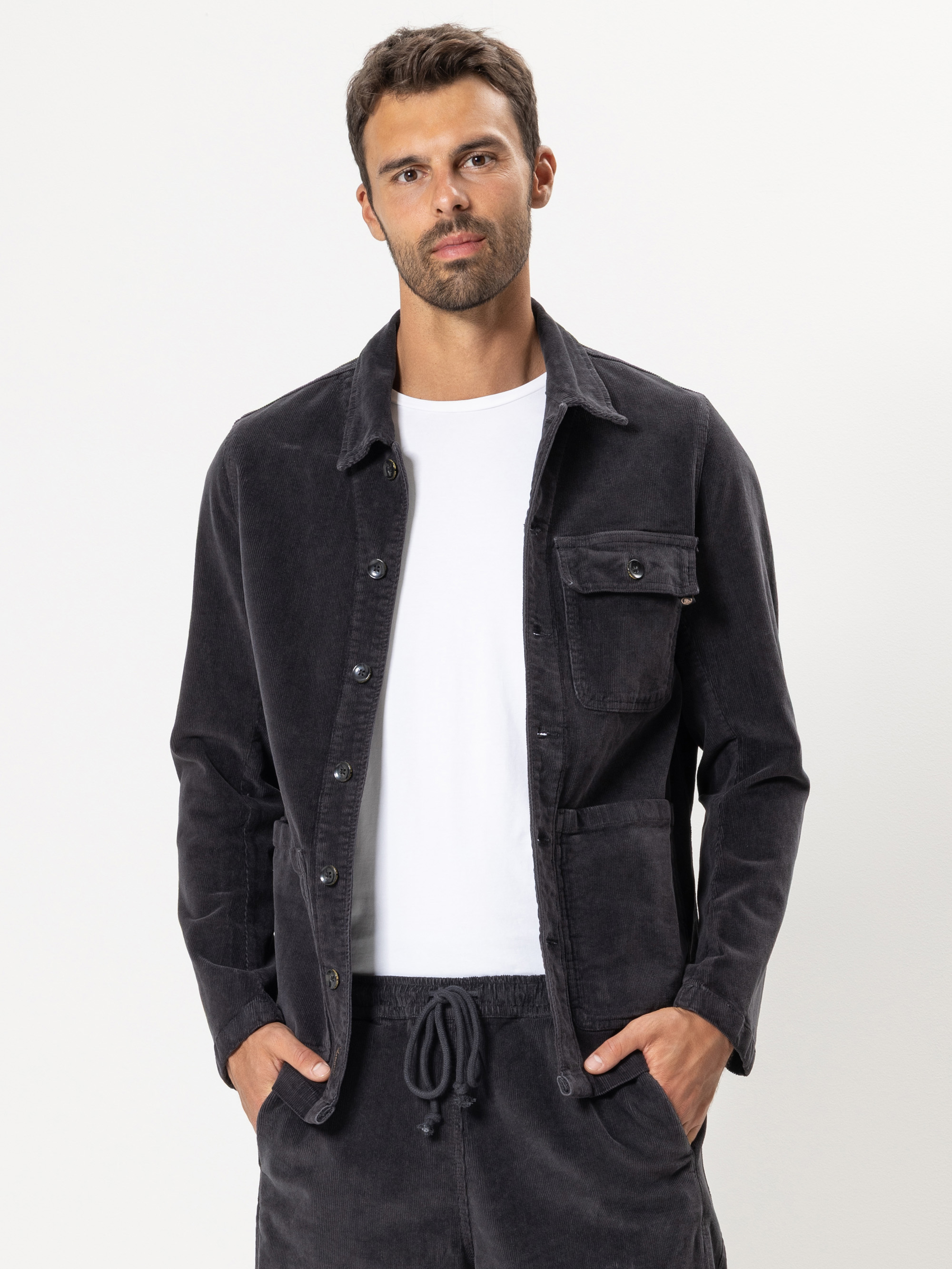 Ein Mann mit kurzen braunen Haaren und einem Bart trägt eine schwarze Cordjacke über einem weißen T-Shirt und eine dazu passende schwarze Hose. Er steht vor einem schlichten weißen Hintergrund und hat die Hände in den Taschen.