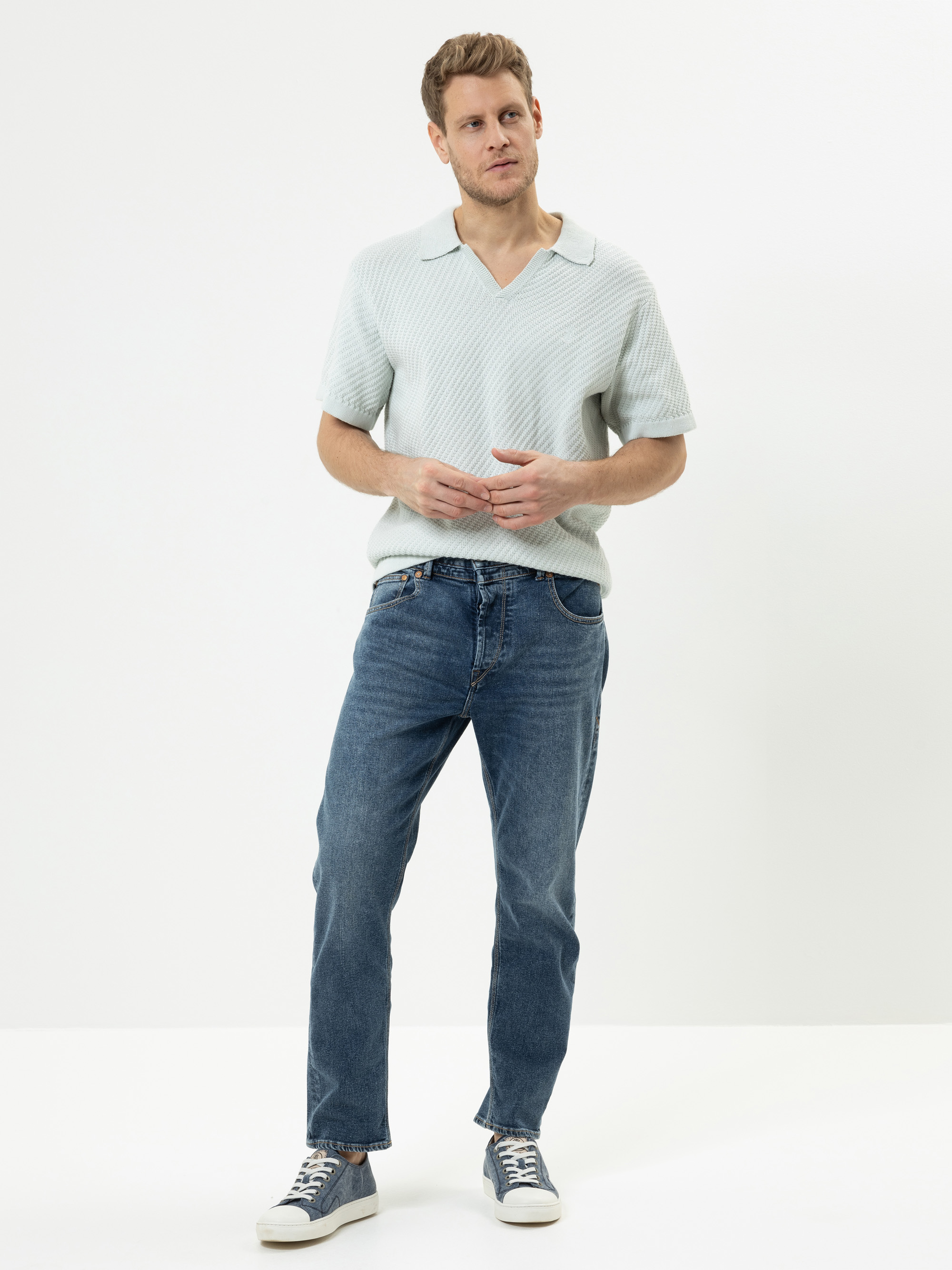 Ein Mann steht vor einem weißen Hintergrund und trägt ein helles kurzärmeliges Hemd mit Kragen, blaue Jeans und graue Turnschuhe. Er hat helles Haar und einen gestutzten Bart, und seine Hände sind locker vor ihm verschränkt.