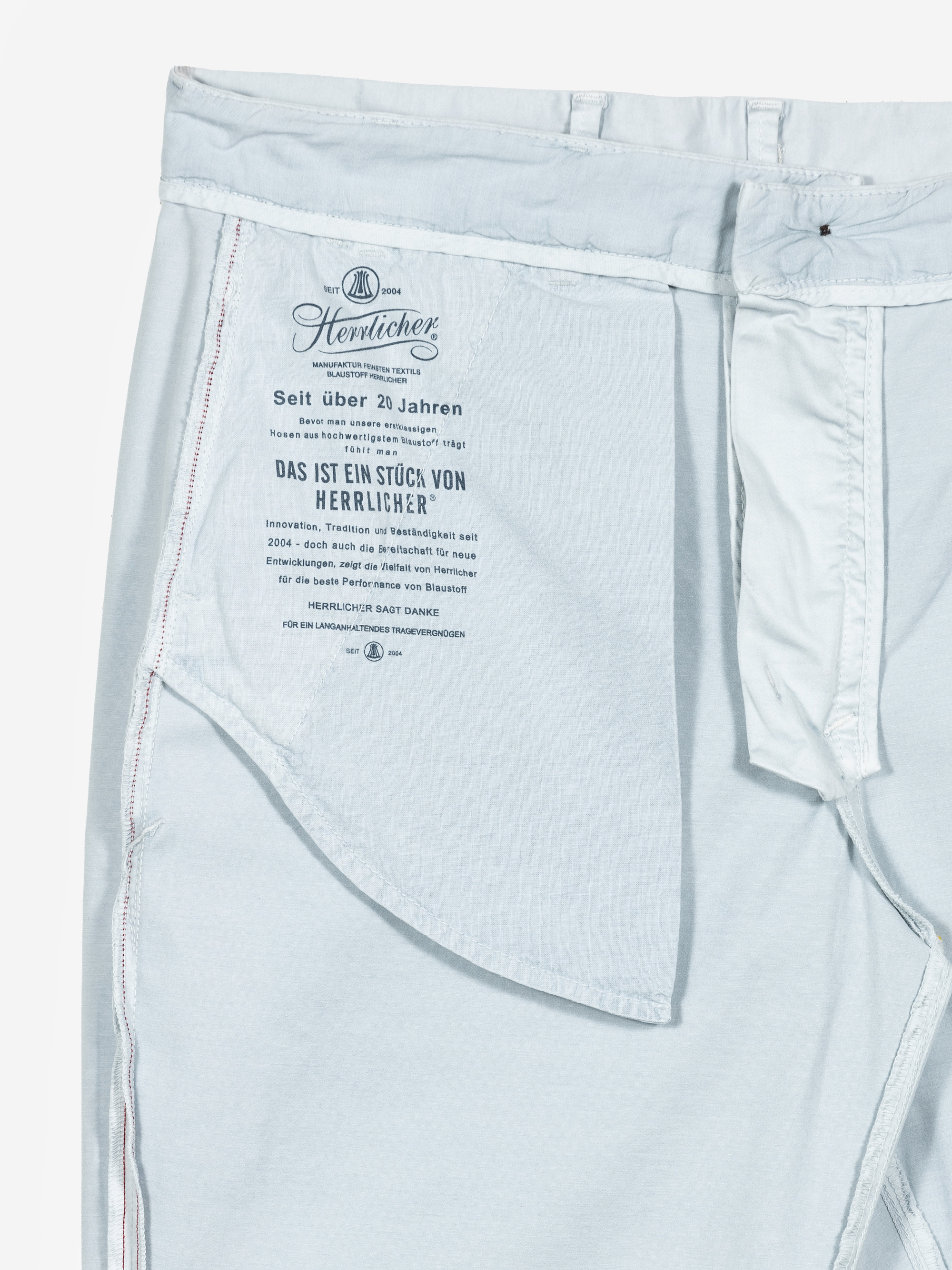 Hellblaue Jeans auf links gedreht, mit sichtbaren Nähten, Taschenfutter und einem gedruckten Etikett mit deutschem Text und Branding-Details von Herrlicher. Der Bund und der Hosenschlitz sind sichtbar.