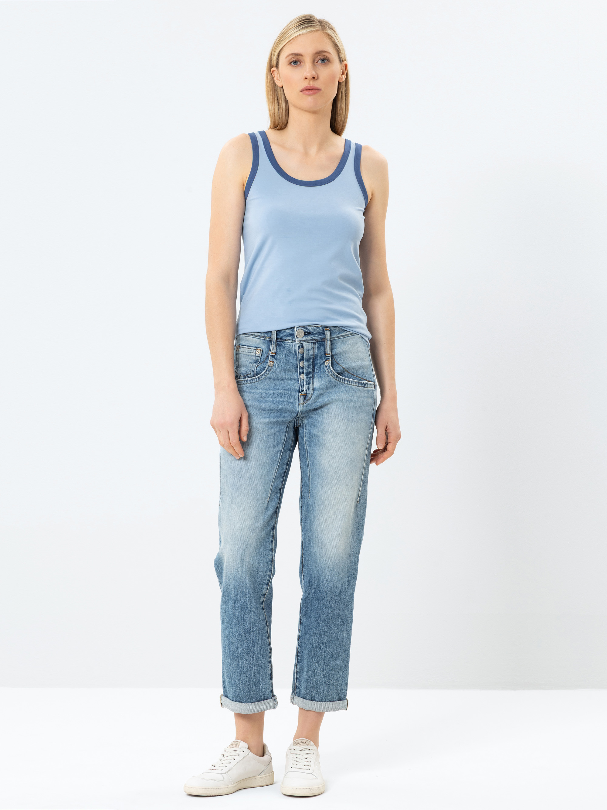 Eine Frau steht vor einem schlichten Hintergrund und trägt ein hellblaues, ärmelloses Tanktop, hellblaue Jeans mit aufgerollten Bündchen und weiße Turnschuhe. Sie hat glattes blondes Haar und einen neutralen Gesichtsausdruck.