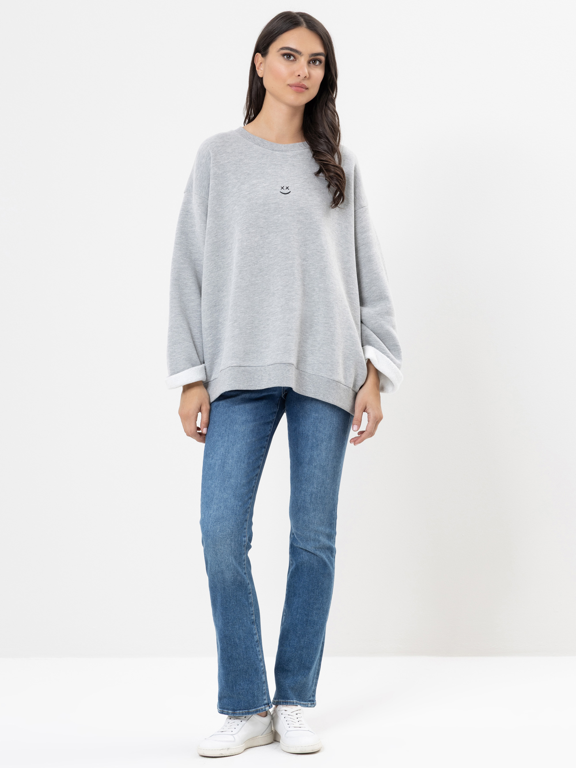 Eine Person steht vor einem schlichten weißen Hintergrund und trägt ein lockeres graues Sweatshirt mit einem kleinen Logo, blaue Jeans und weiße Turnschuhe. Sie hat lange dunkle Haare und einen neutralen Gesichtsausdruck.