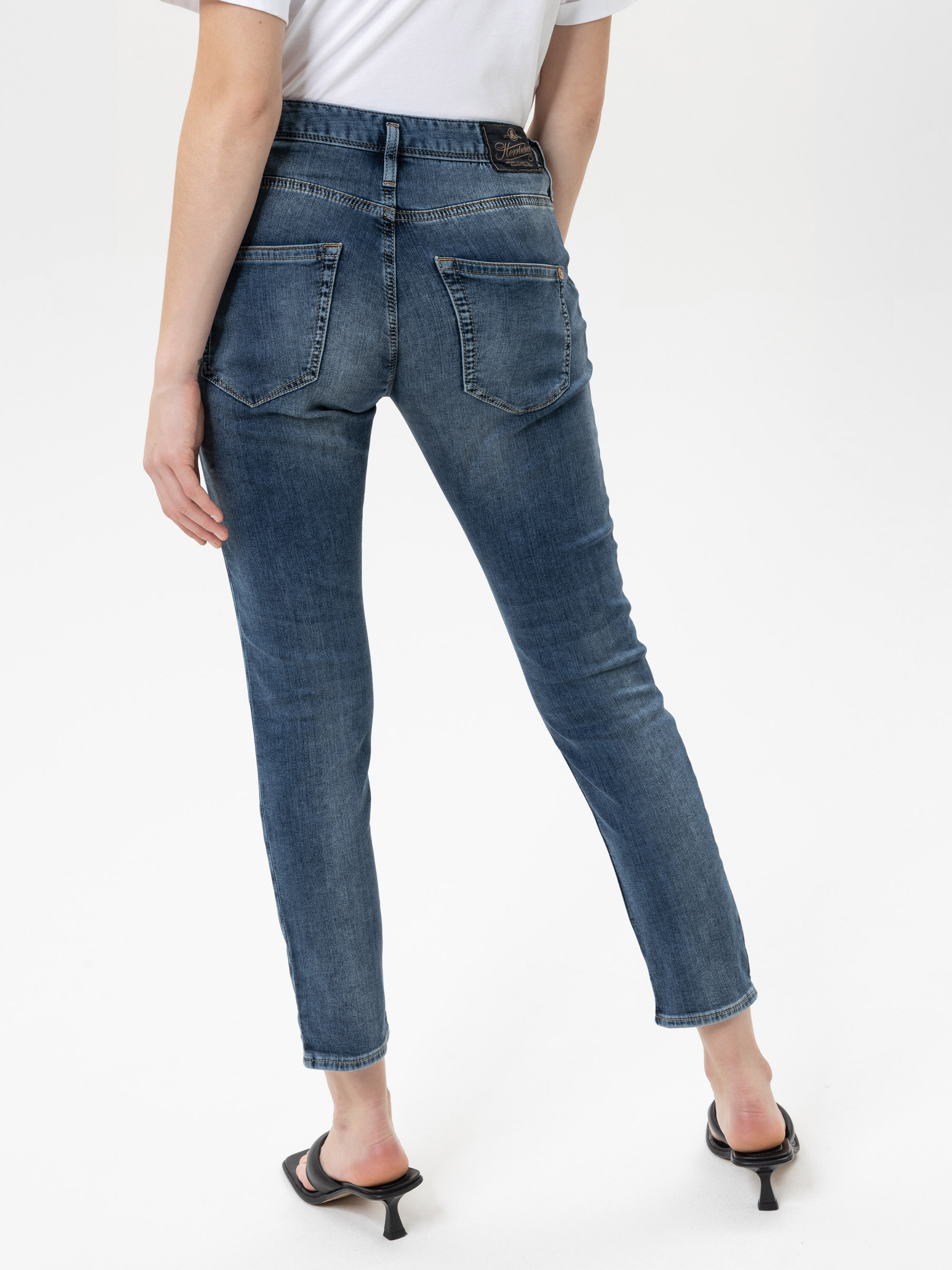 Eine Person, die ein weißes Hemd, blaue Röhrenjeans und schwarze hochhackige Sandalen trägt, steht mit dem Rücken zur Kamera vor einem schlichten weißen Hintergrund.