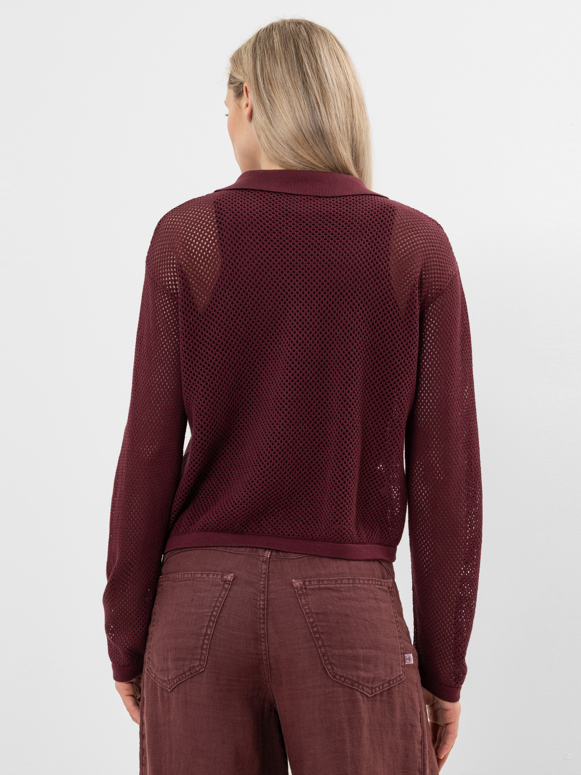 Eine Frau mit langen blonden Haaren ist von hinten zu sehen. Sie trägt ein burgunderrotes langärmeliges Netzoberteil und eine dazu passende hochtaillierte Hose. Sie steht vor einem schlichten, hellen Hintergrund.