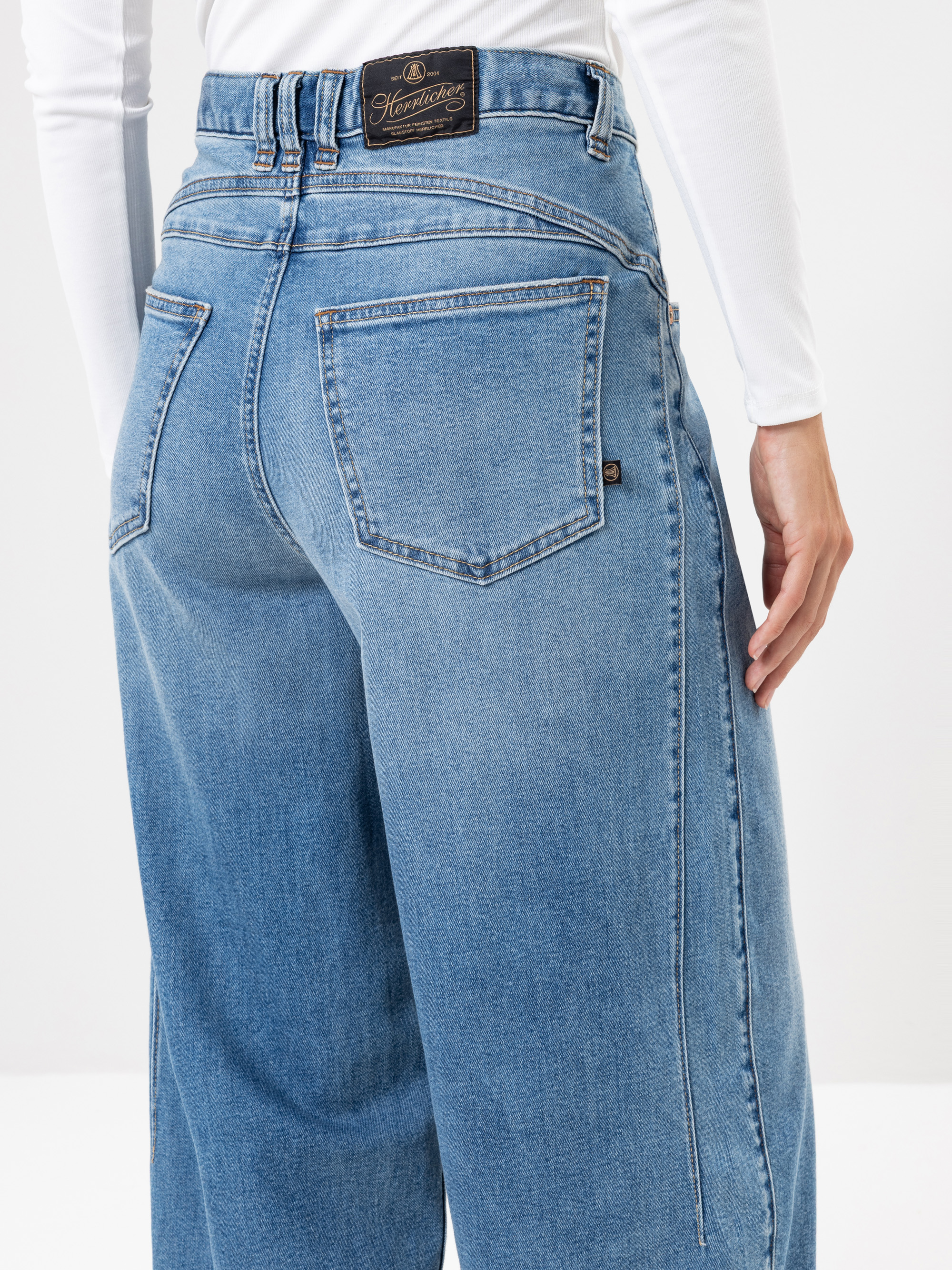 Eine Person, die eine hoch taillierte, locker sitzende blaue Jeans und ein weißes langärmeliges Oberteil trägt, von hinten gesehen, wobei die beiden Gesäßtaschen der Jeans und ein Markenetikett am Bund hervorgehoben werden.