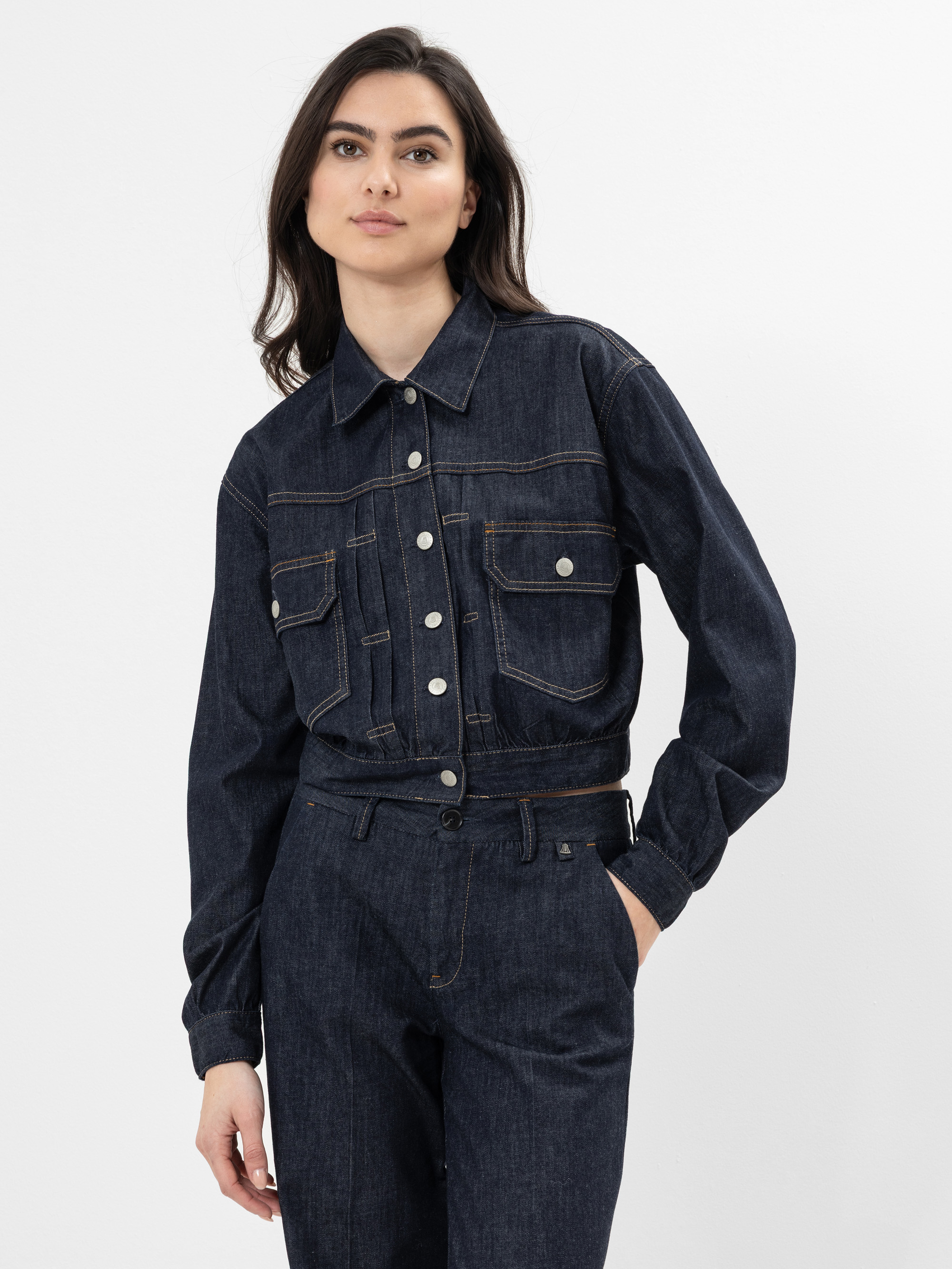 Eine Frau mit langen dunklen Haaren trägt eine dunkle Jeans-Knopfjacke mit Vordertaschen und eine dazu passende Jeans mit hoher Taille. Sie steht vor einem schlichten weißen Hintergrund.