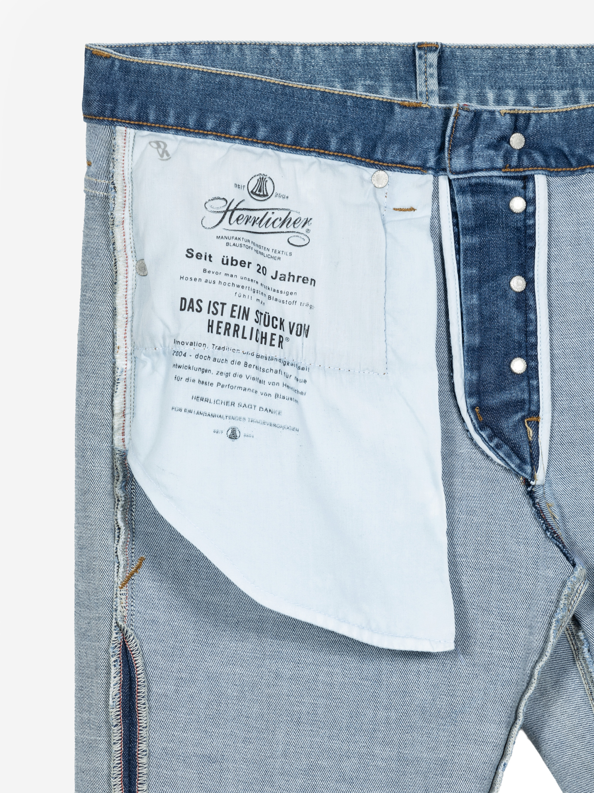 Innenansicht einer blauen Jeans mit Schwerpunkt auf der Innentasche, die einen Markentext und Produktinformationen in deutscher Sprache enthält. Die Jeans hat sichtbare Nähte und silberne Nietenknöpfe am Bund und Hosenschlitz.