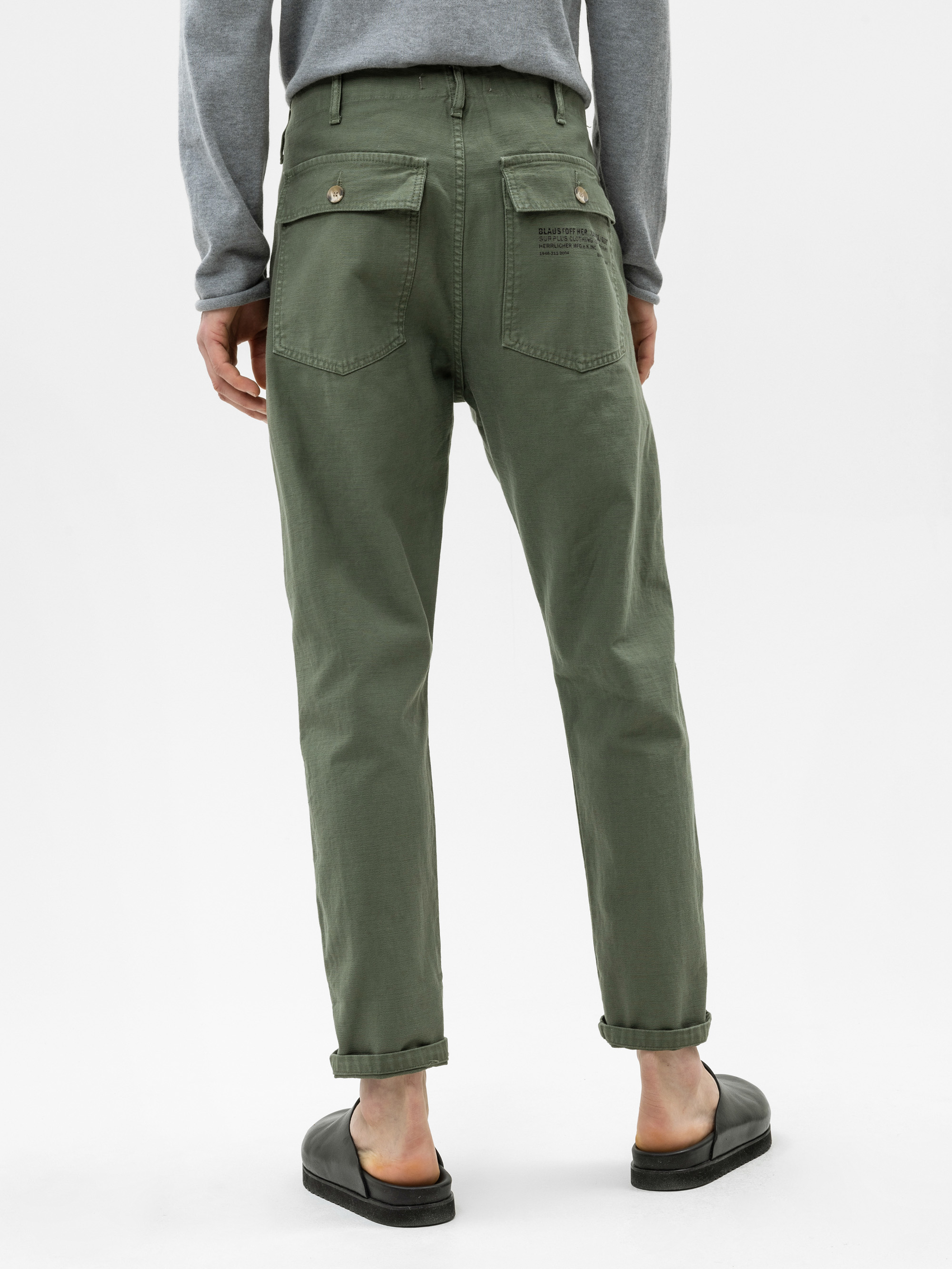 Eine Person, die eine olivgrüne Hose mit Bündchen am Saum, Gesäßtaschen mit Klappe und Knöpfen, ein graues langärmeliges Hemd und schwarze Slide-Sandalen trägt, steht vor einem schlichten weißen Hintergrund.