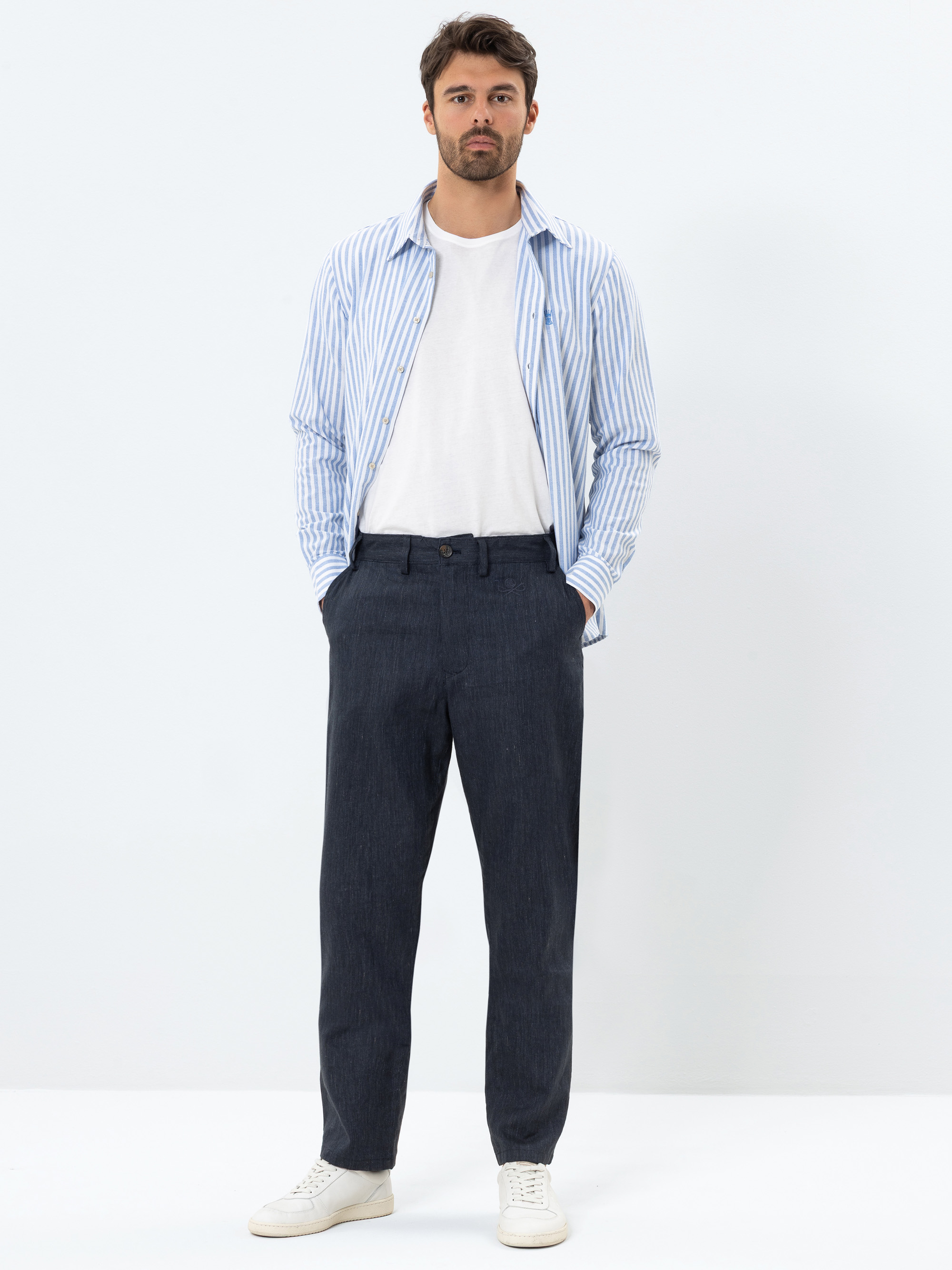 Ein Mann steht vor einem schlichten weißen Hintergrund, trägt ein blau-weiß gestreiftes Hemd über einem weißen T-Shirt, eine dunkle Hose und weiße Turnschuhe, die Hände in den Taschen.