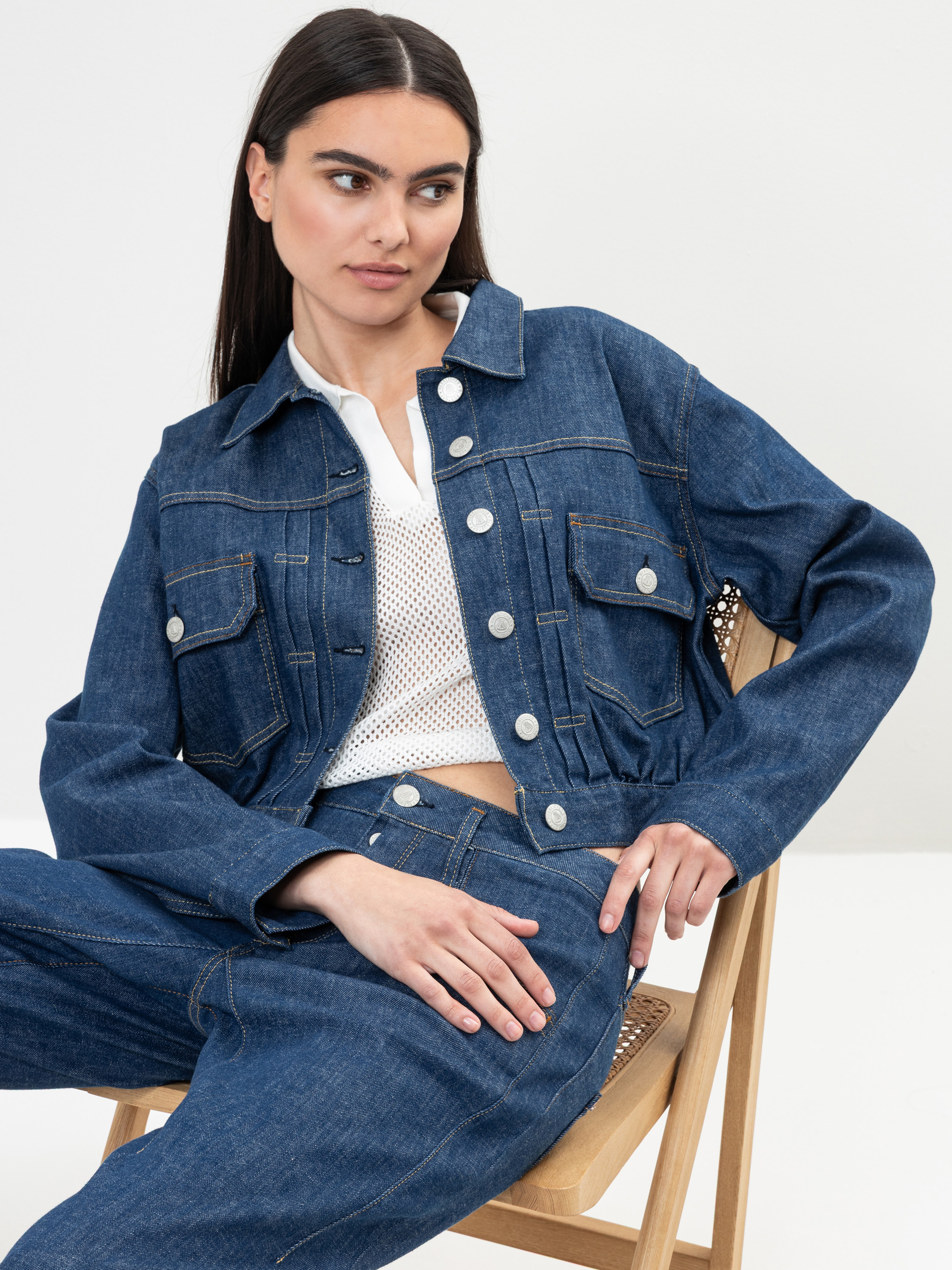 Eine Frau mit langen dunklen Haaren sitzt auf einem Stuhl und trägt eine blaue Jeansjacke und passende Jeans über einem weißen Netzoberteil. Sie blickt mit neutraler Miene vor einem schlichten Hintergrund zur Seite.