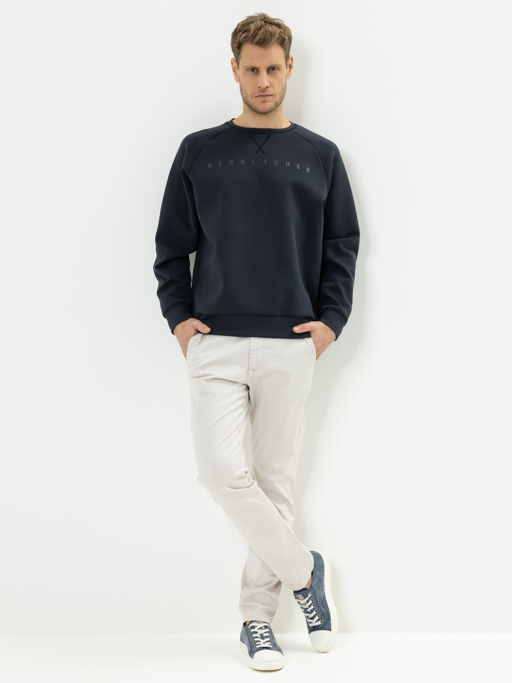 Ein Mann steht vor einem schlichten weißen Hintergrund und trägt ein marineblaues Sweatshirt, eine hellbeige Hose und blaue Turnschuhe. Er hat die Hände in den Taschen und ein Bein über das andere geschlagen.