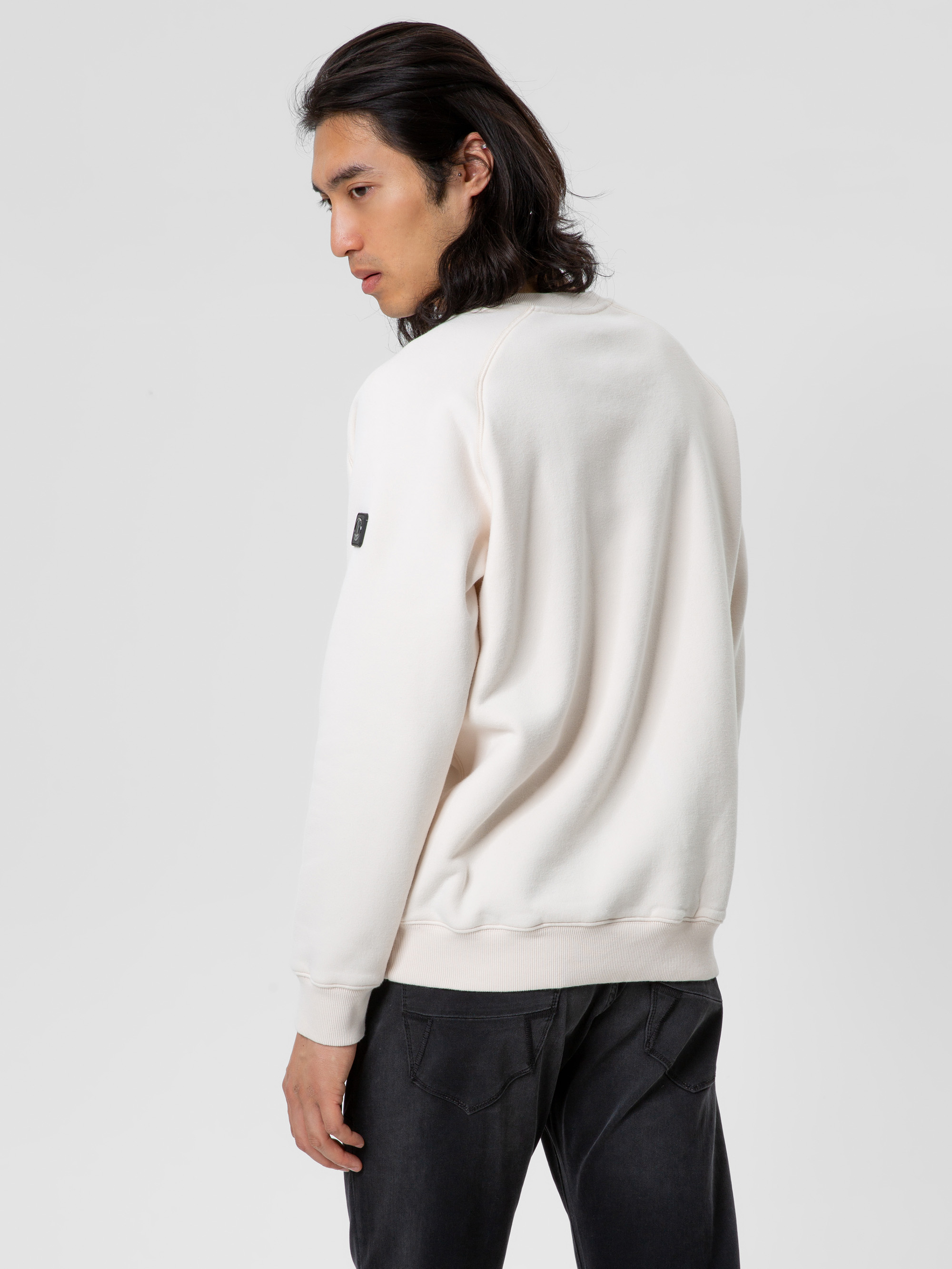 Eine Person mit langen dunklen Haaren, bekleidet mit einem hellen Sweatshirt und einer schwarzen Jeans, steht und schaut über ihre linke Schulter vor einem schlichten hellen Hintergrund.