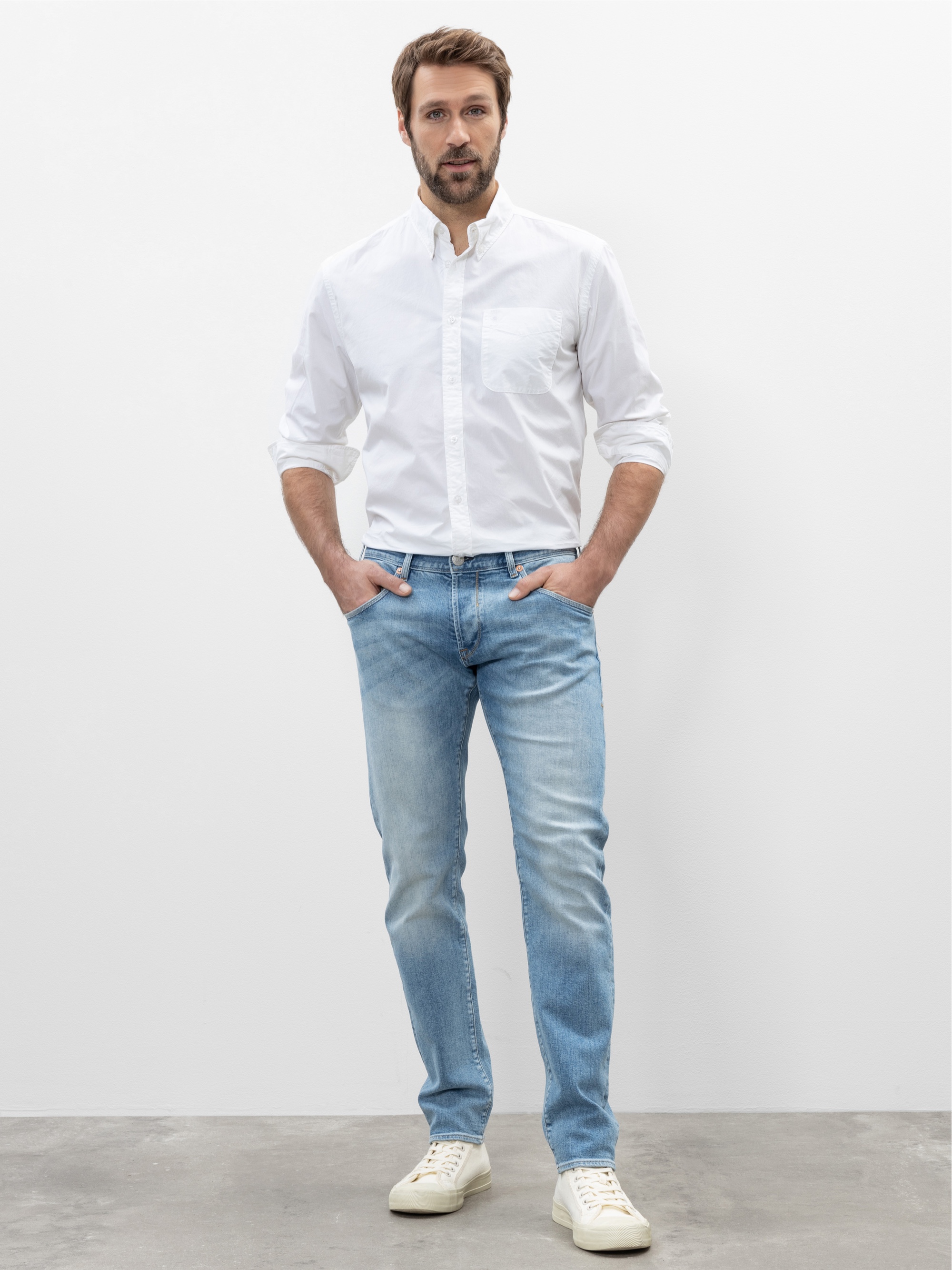 Ein Mann mit kurzen braunen Haaren und Bart steht vor einem schlichten weißen Hintergrund. Er trägt ein weißes Button-down-Hemd, hellblaue Jeans und weiße Turnschuhe und hat die Hände in den Taschen.