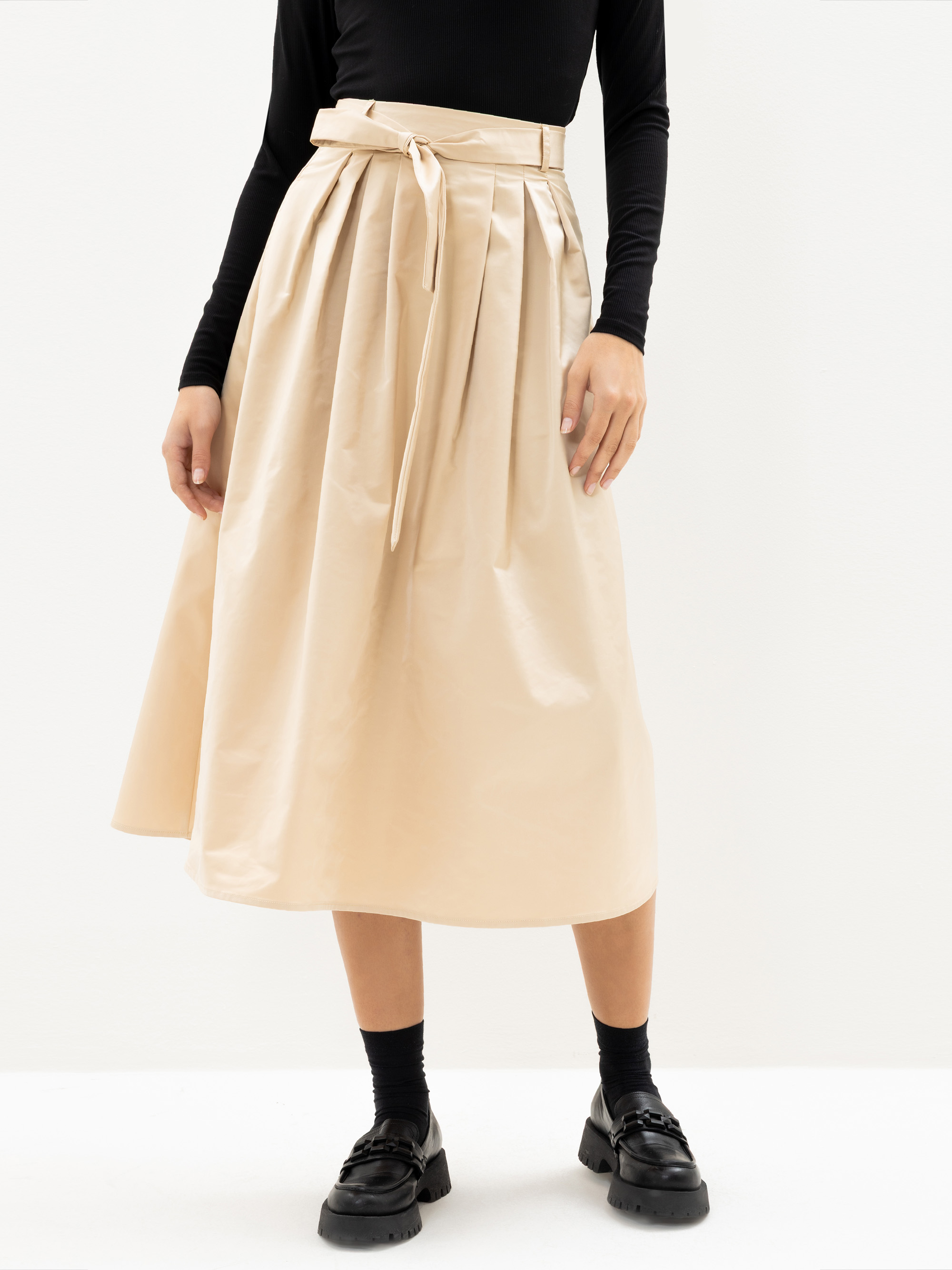Eine Person, die einen beigefarbenen Midirock mit hoher Taille, Falten und einem passenden Gürtel trägt, dazu ein schwarzes langärmeliges Oberteil, schwarze Socken und schwarze Slipper, vor einem schlichten weißen Hintergrund.