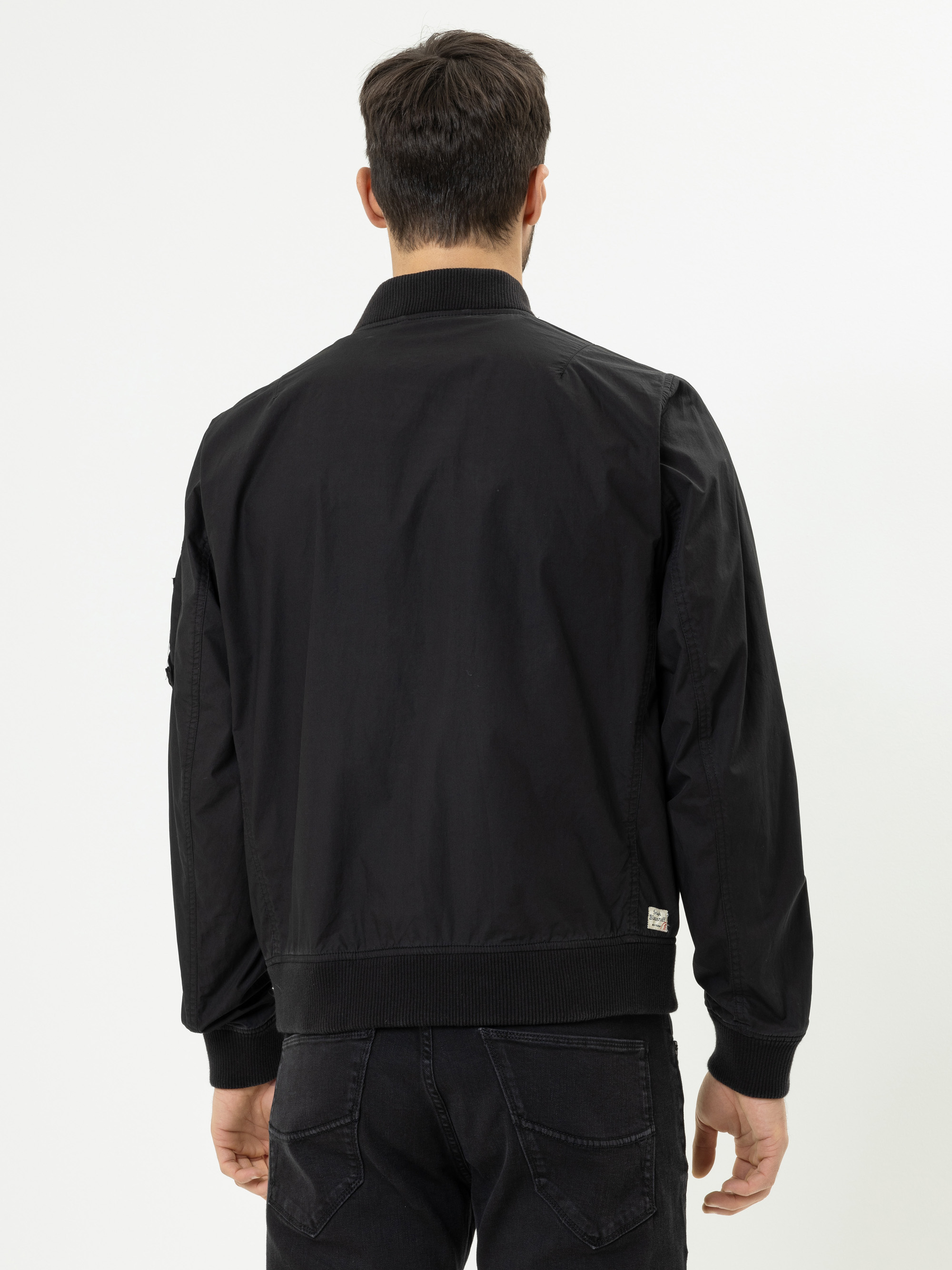 Ein Mann mit kurzen dunklen Haaren wird von hinten gezeigt. Er trägt eine schwarze Bomberjacke und schwarze Jeans vor einem schlichten weißen Hintergrund.