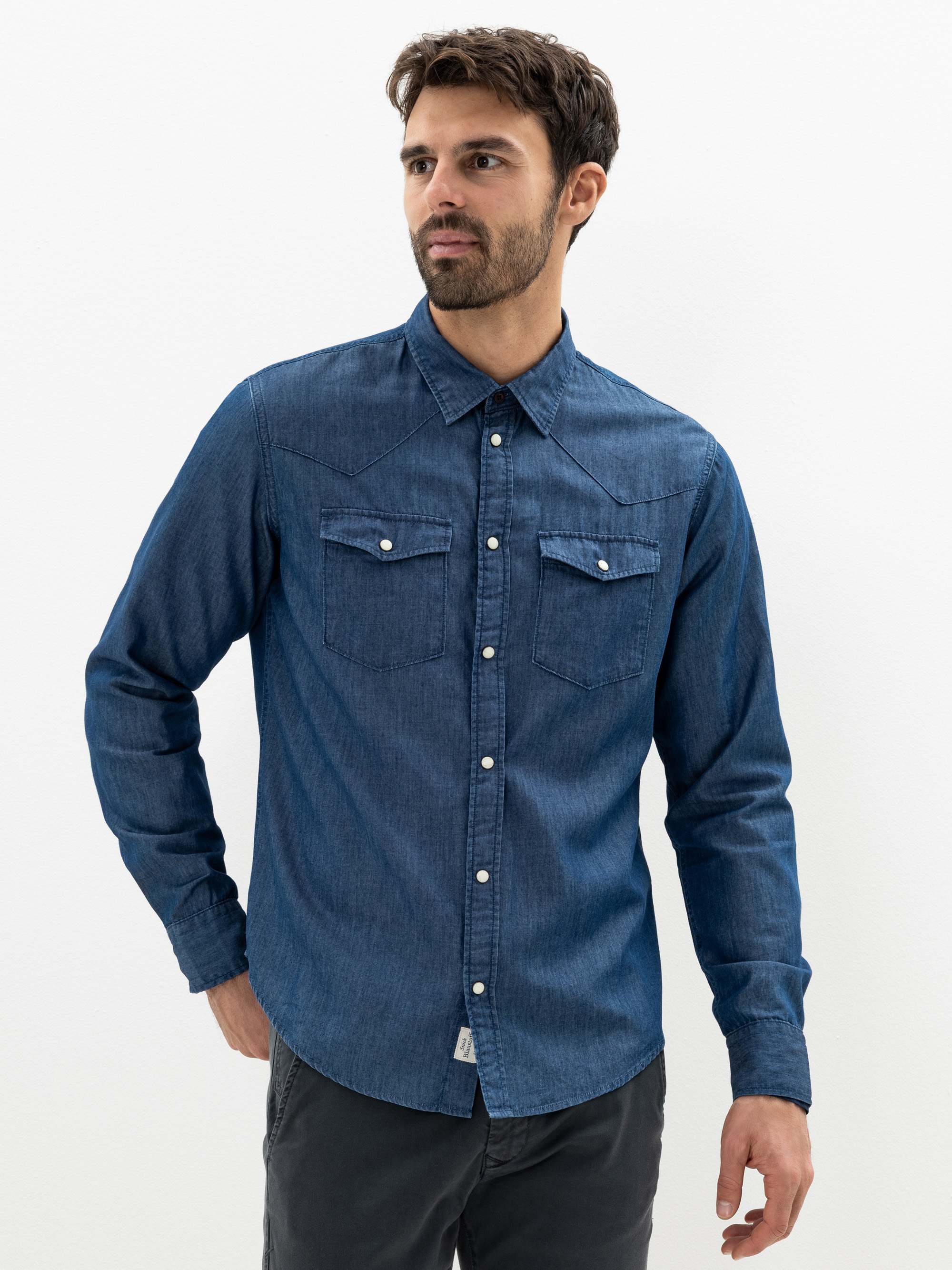 Ein Mann mit kurzen braunen Haaren und einem Bart trägt ein langärmeliges, hochgeknöpftes Jeanshemd mit zwei Vordertaschen und eine dunkle Hose und steht vor einem schlichten weißen Hintergrund.