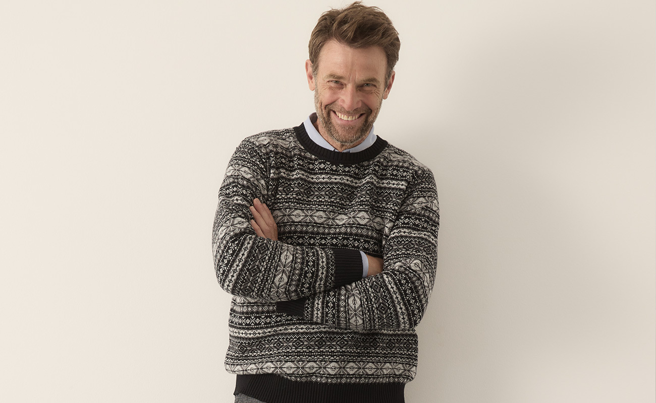 Ein Mann mit kurzen braunen Haaren und einem Bart lächelt mit verschränkten Armen. Er trägt einen gemusterten schwarz-weiß-grauen Pullover und steht vor einem schlichten hellen Hintergrund.