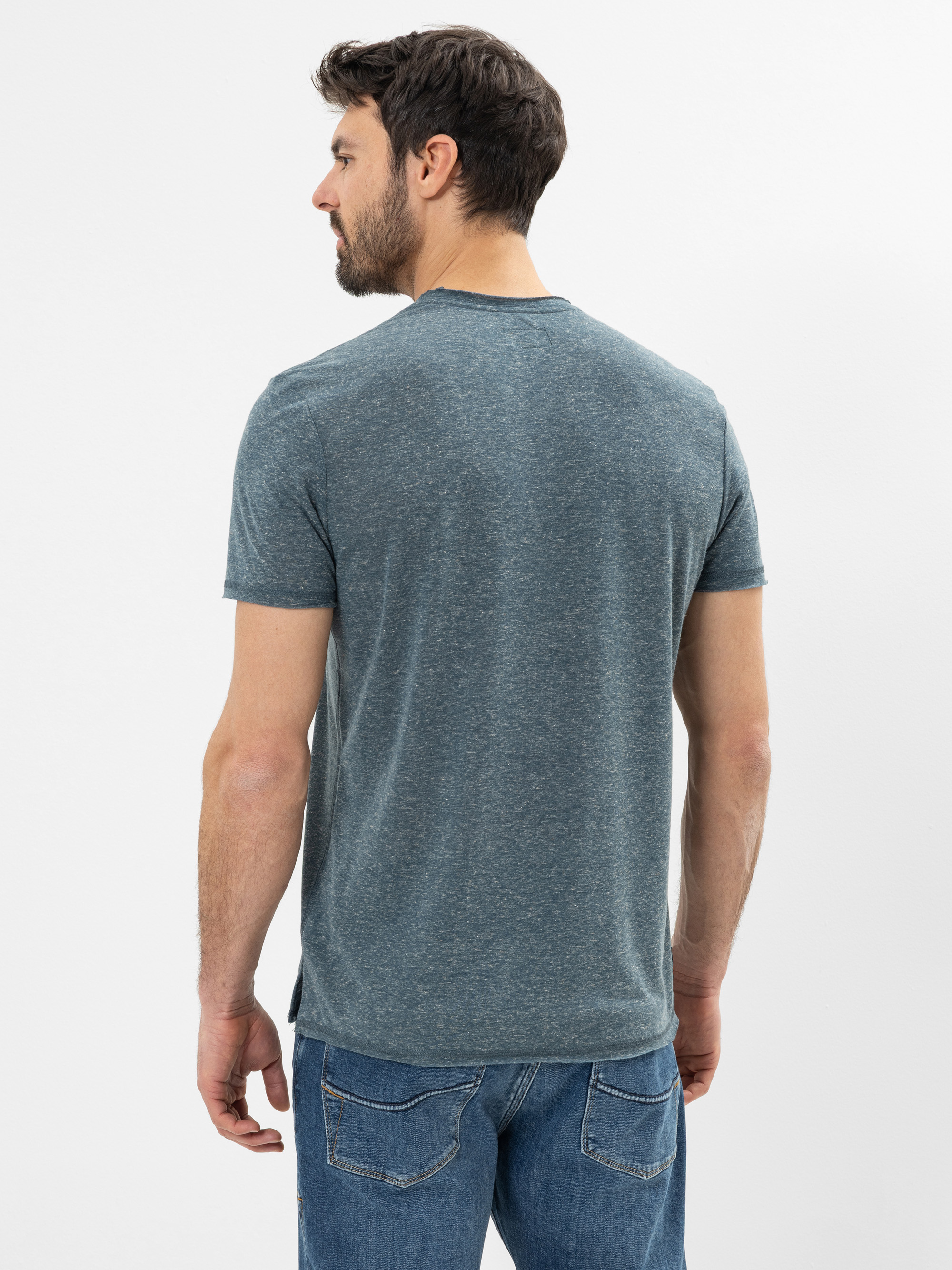 Ein Mann mit kurzen dunklen Haaren und einem Bart steht und schaut von der Kamera weg. Er trägt ein beheiztes blaues T-Shirt und eine blaue Jeans vor einem schlichten, hellen Hintergrund.