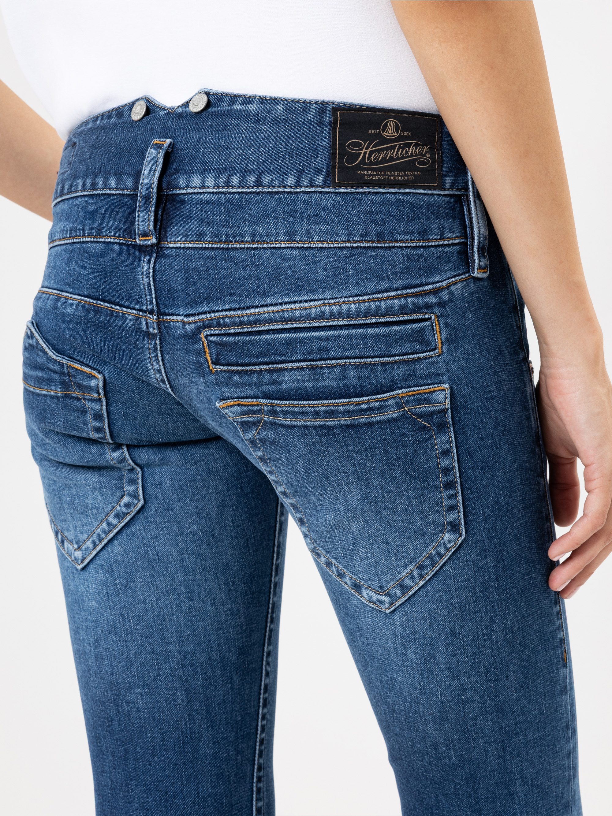 Eine Person, die eine blaue Jeans und ein weißes Oberteil trägt, wird von der Taille abwärts gezeigt, wobei die Gesäßtaschen und ein schwarzer Markenaufnäher am Hosenbund hervorgehoben werden.
