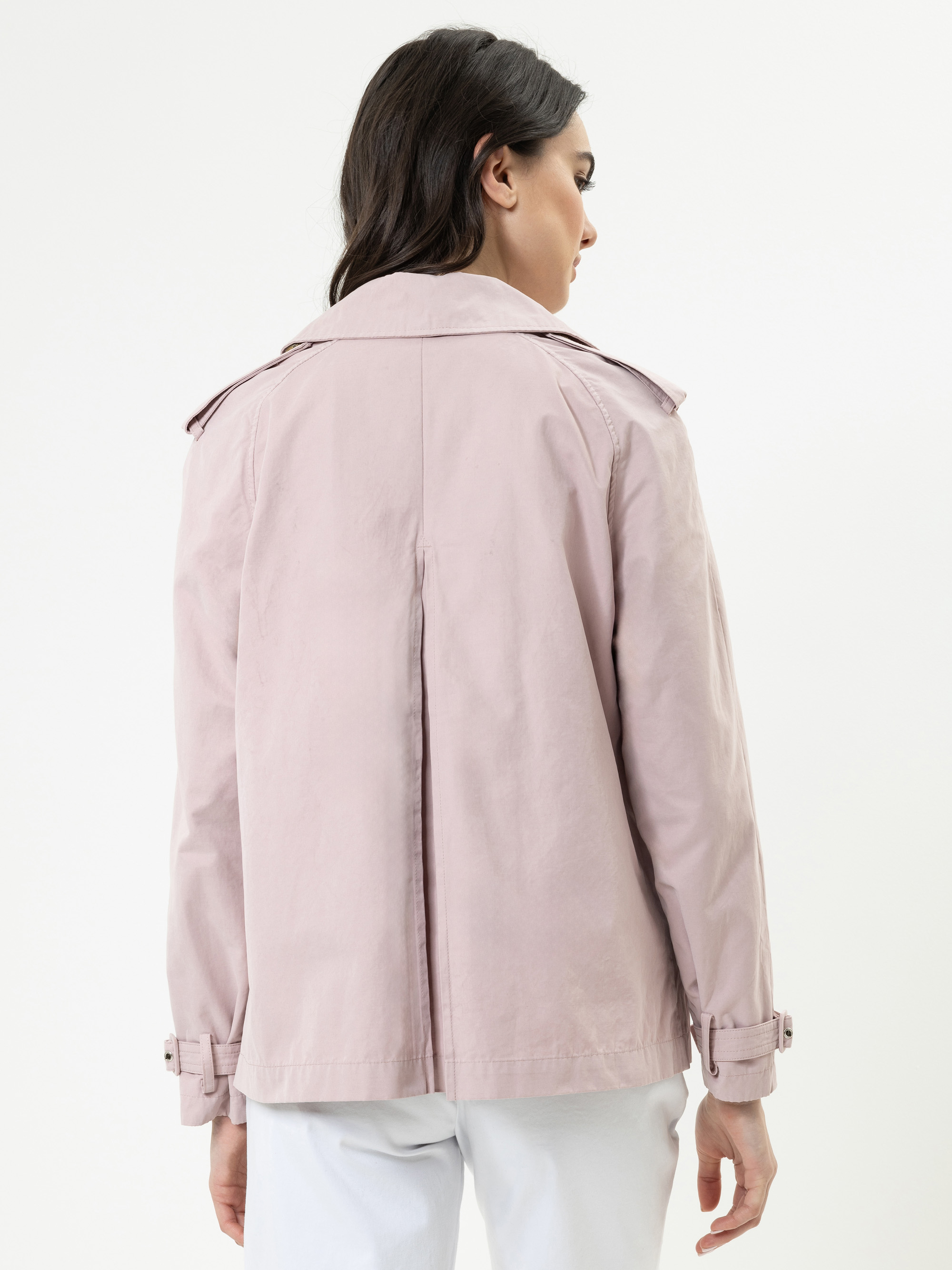 Eine Frau steht mit dem Gesicht nach hinten und trägt eine hellrosa Jacke mit Schulterklappen und Schnallen an den Ärmeln. Sie hat dunkles Haar und trägt ebenfalls eine weiße Hose. Der Hintergrund ist einfarbig weiß.