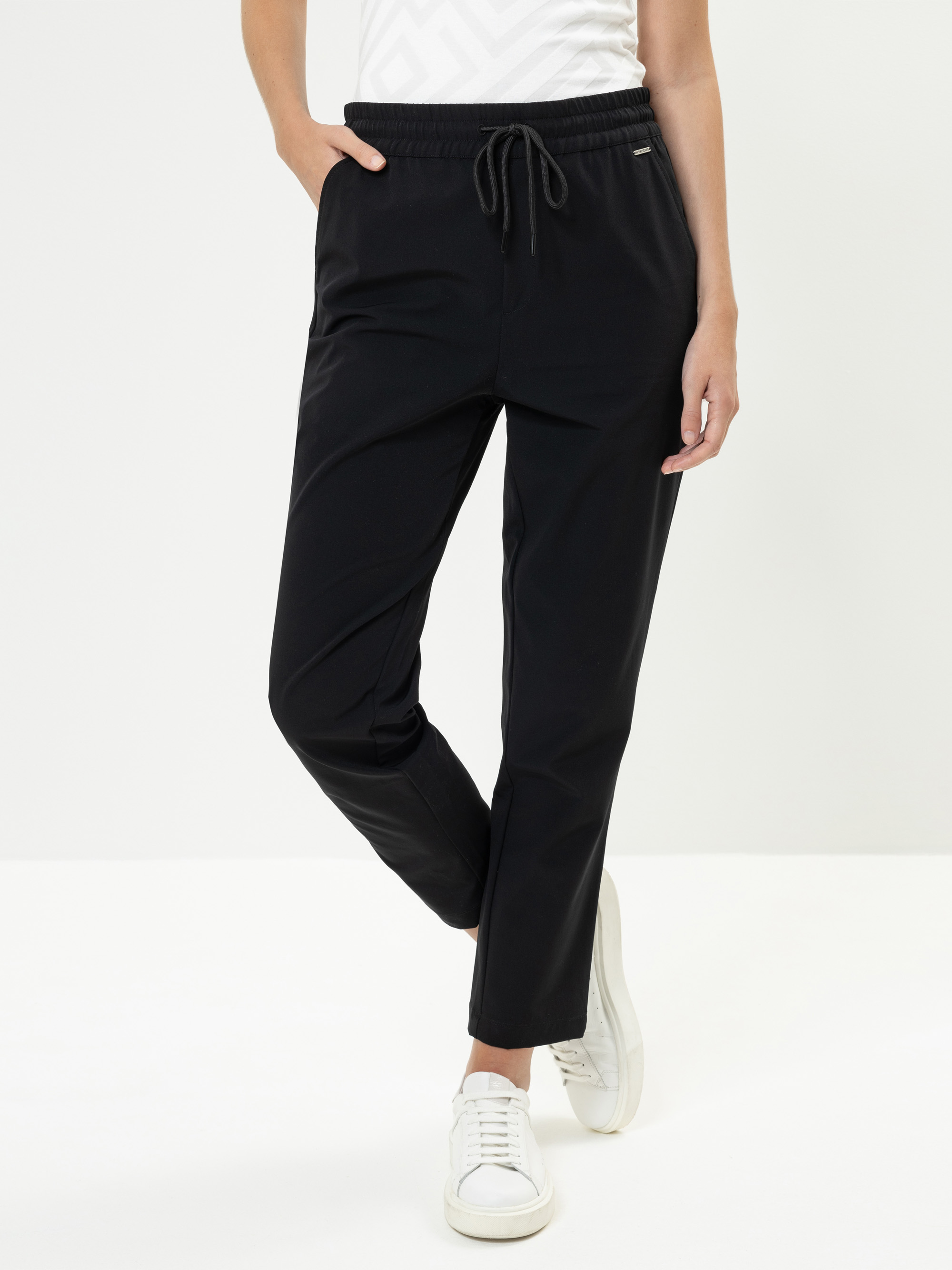 Eine Person, die eine schwarze Hose mit Kordelzug, ein weißes gemustertes Hemd und weiße Turnschuhe trägt, steht mit den Händen in den Taschen vor einem schlichten weißen Hintergrund.
