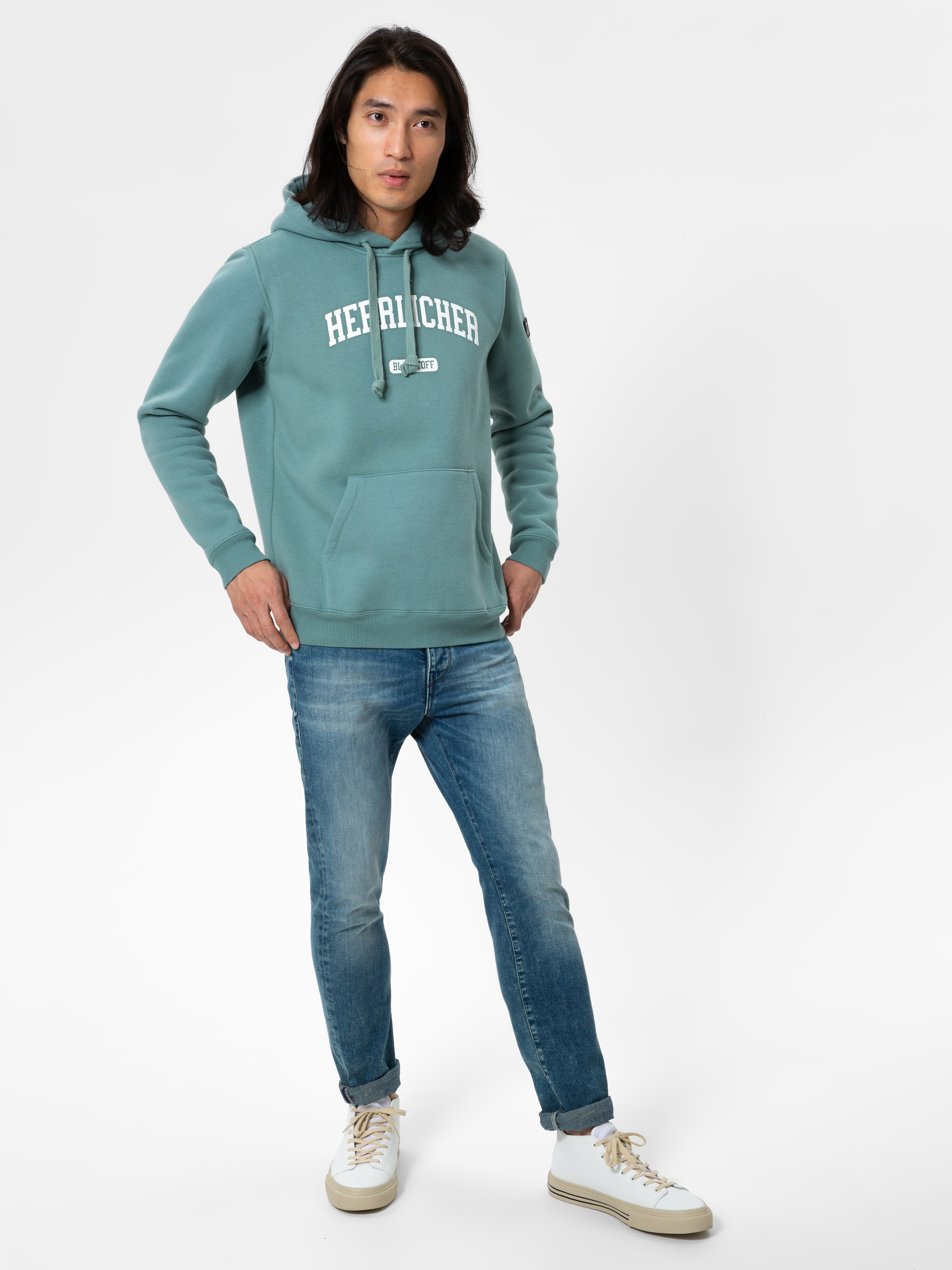 Eine Person mit langen dunklen Haaren trägt ein blaugrünes Kapuzensweatshirt mit der Aufschrift HEFRICHER, blaue Jeans und weiße Turnschuhe und steht vor einem schlichten weißen Hintergrund.