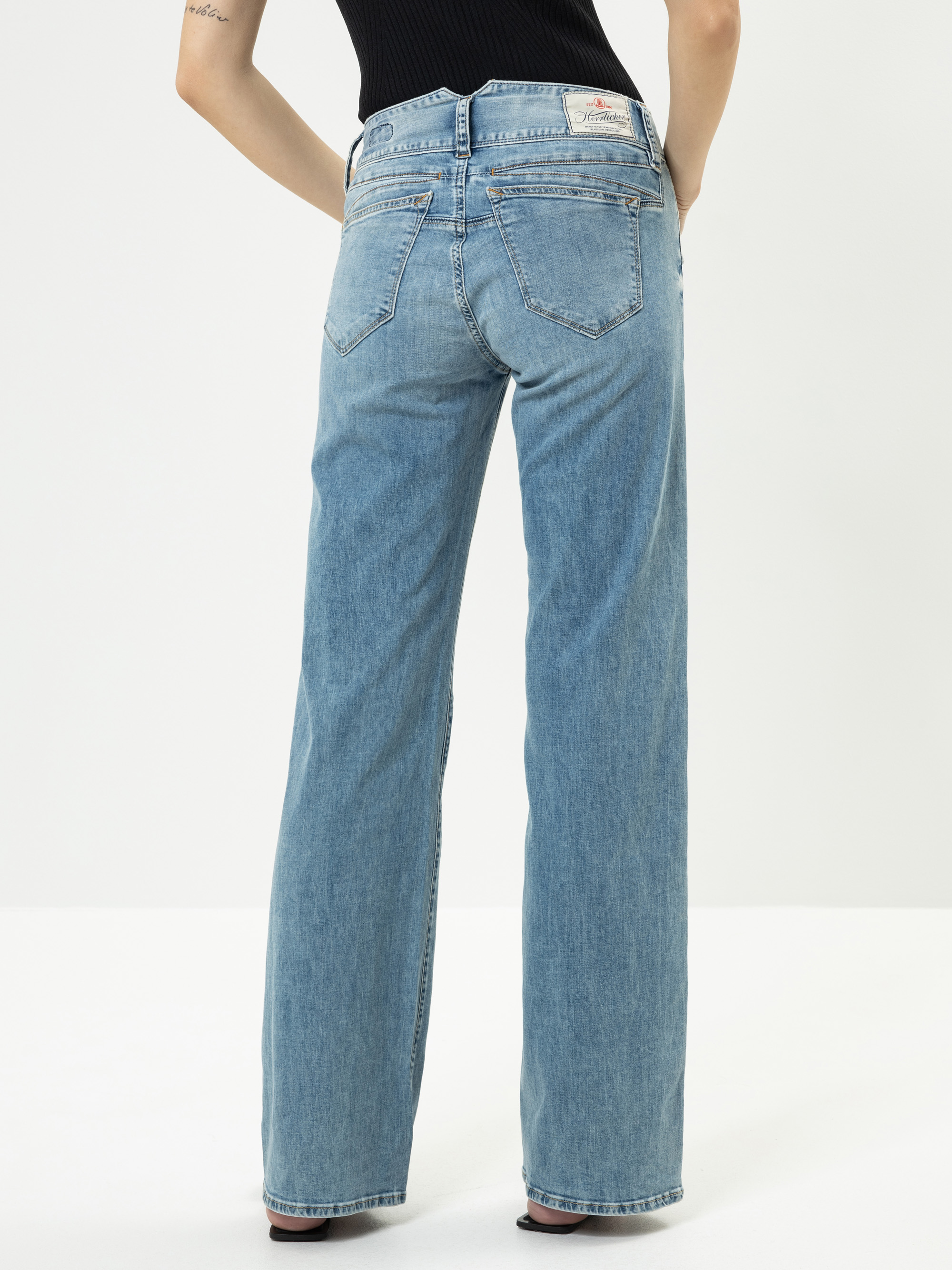 Die Person steht mit dem Rücken zur Kamera und trägt eine hellblaue Jeans mit weitem Bein und ein schwarzes ärmelloses Oberteil. Die Jeans hat ein weißes Etikett am Bund. Der Hintergrund ist einfarbig und weiß.