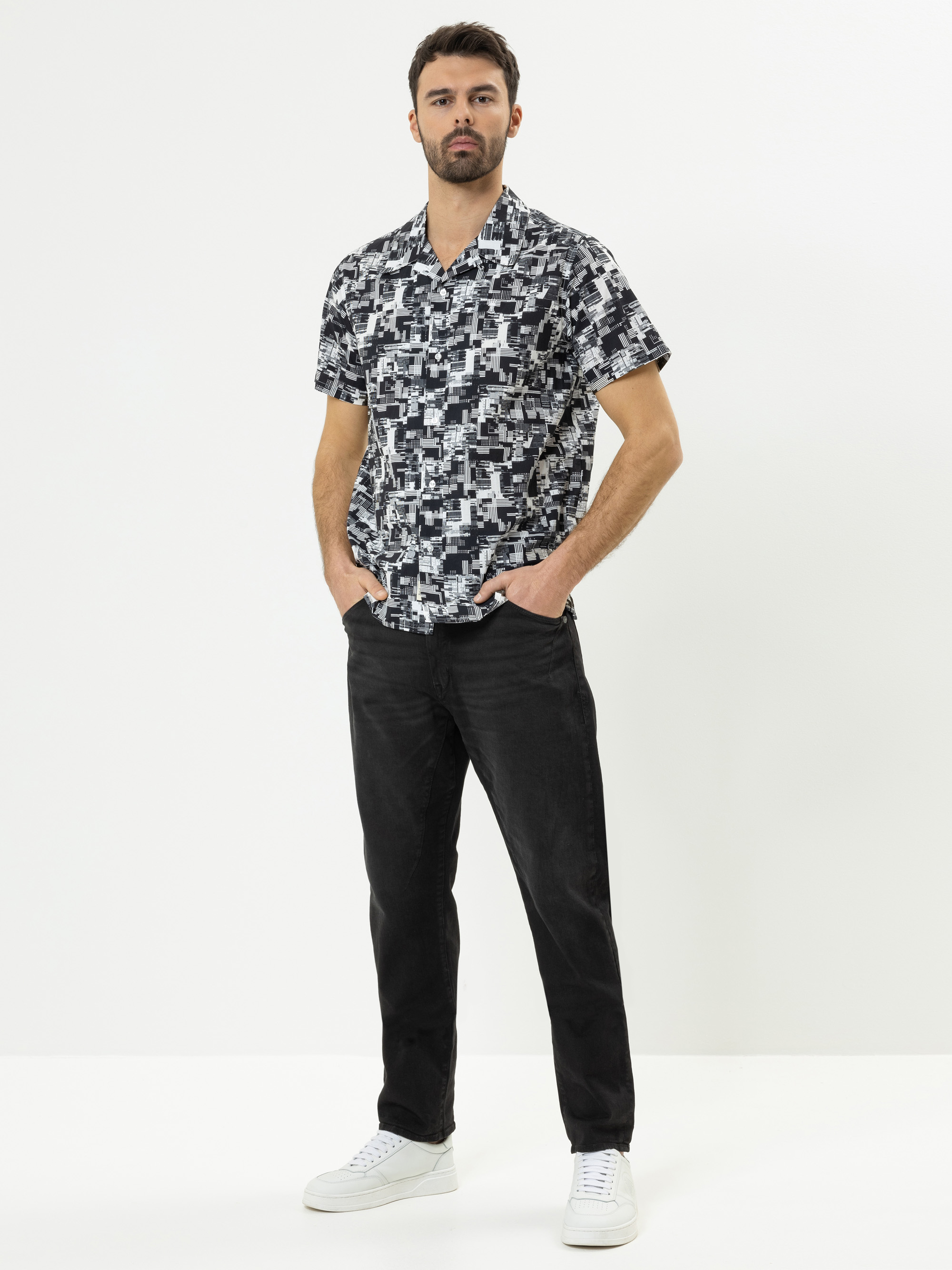 Ein Mann steht vor einem schlichten weißen Hintergrund und trägt ein kurzärmeliges, schwarz-weiß gemustertes Hemd, eine schwarze Hose und weiße Turnschuhe. Er hat die Hände in den Taschen und einen neutralen Gesichtsausdruck.