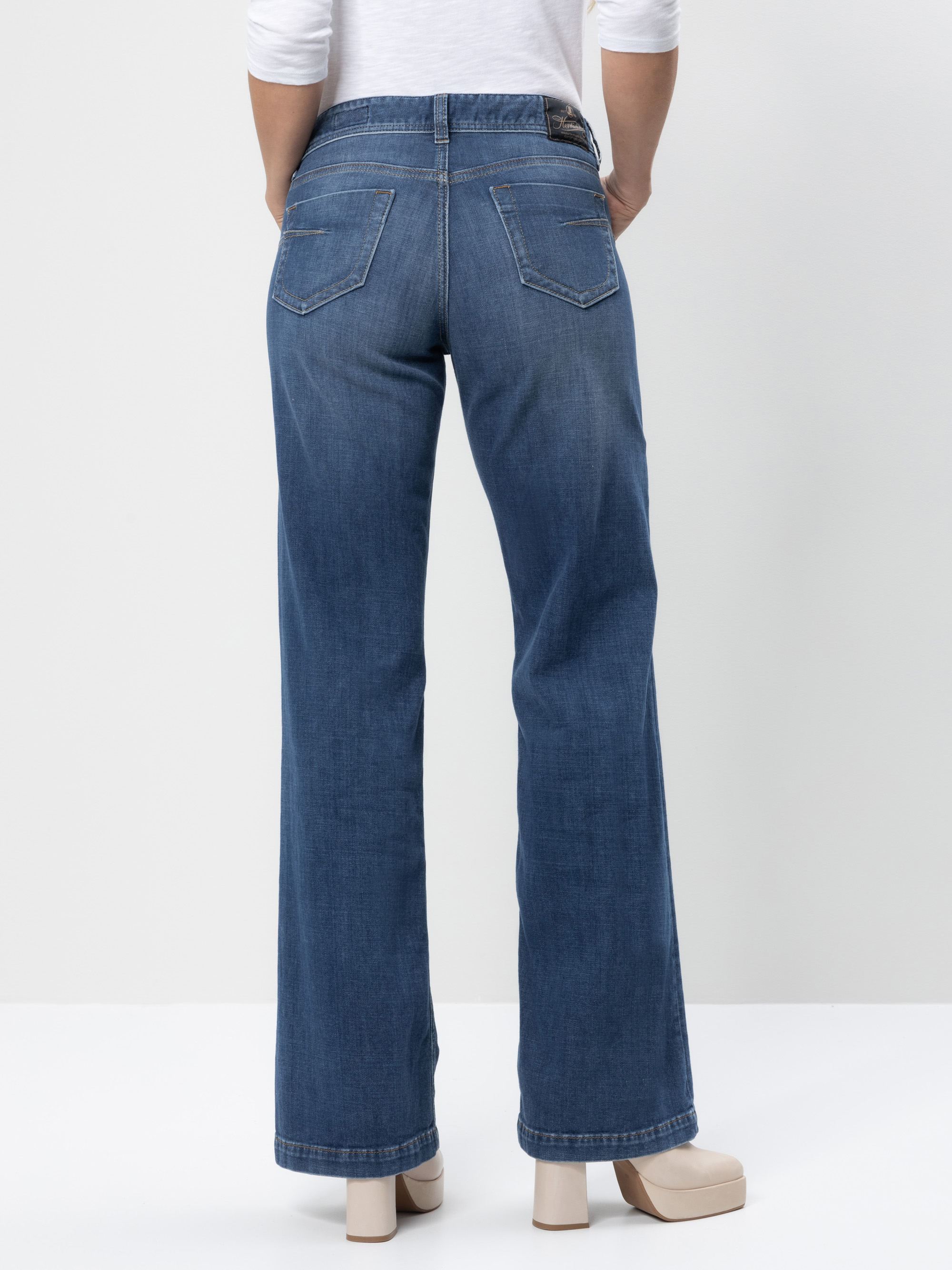 Eine Person, die eine blaue Jeans mit hoher Taille und geradem Bein sowie cremefarbene Plateauschuhe trägt, steht mit dem Gesicht nach außen vor einem schlichten weißen Hintergrund.