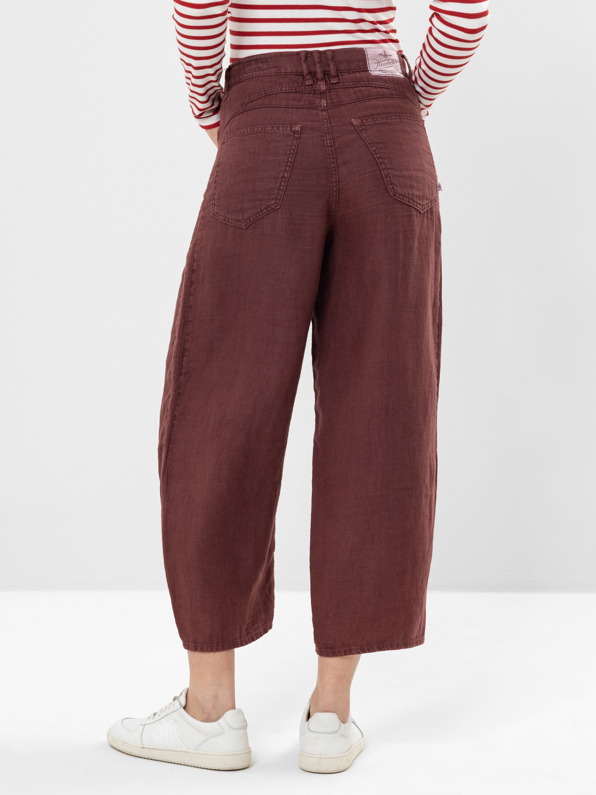 Mit dem Rücken zur Kamera stehende Person, die eine weite kastanienbraune Hose, ein rot-weiß gestreiftes langärmeliges Hemd und weiße Turnschuhe trägt, vor einem schlichten hellen Hintergrund.