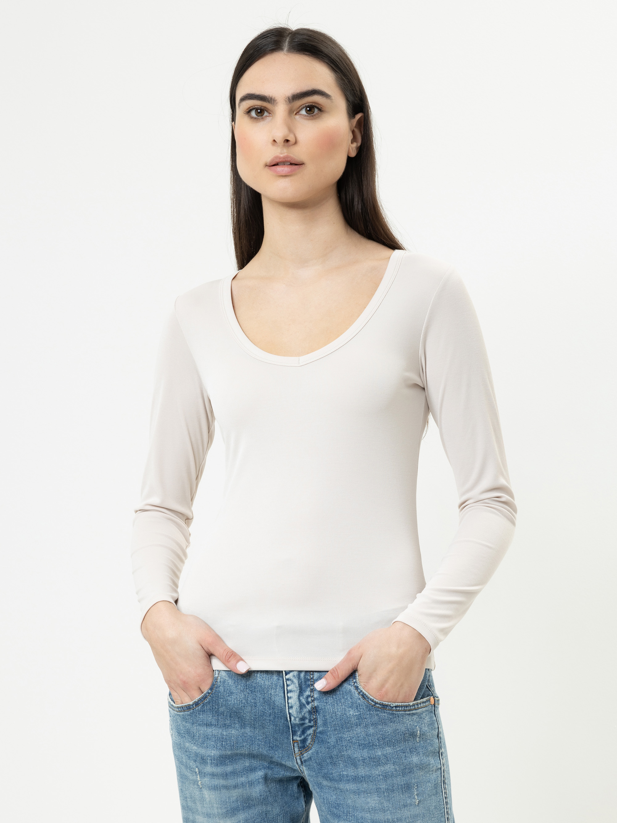 Eine Frau mit langen dunklen Haaren, die ein hellbeiges, langärmeliges Oberteil und blaue Jeans trägt, steht vor einem schlichten weißen Hintergrund und schaut leicht zur Seite, die Hände in den Taschen.