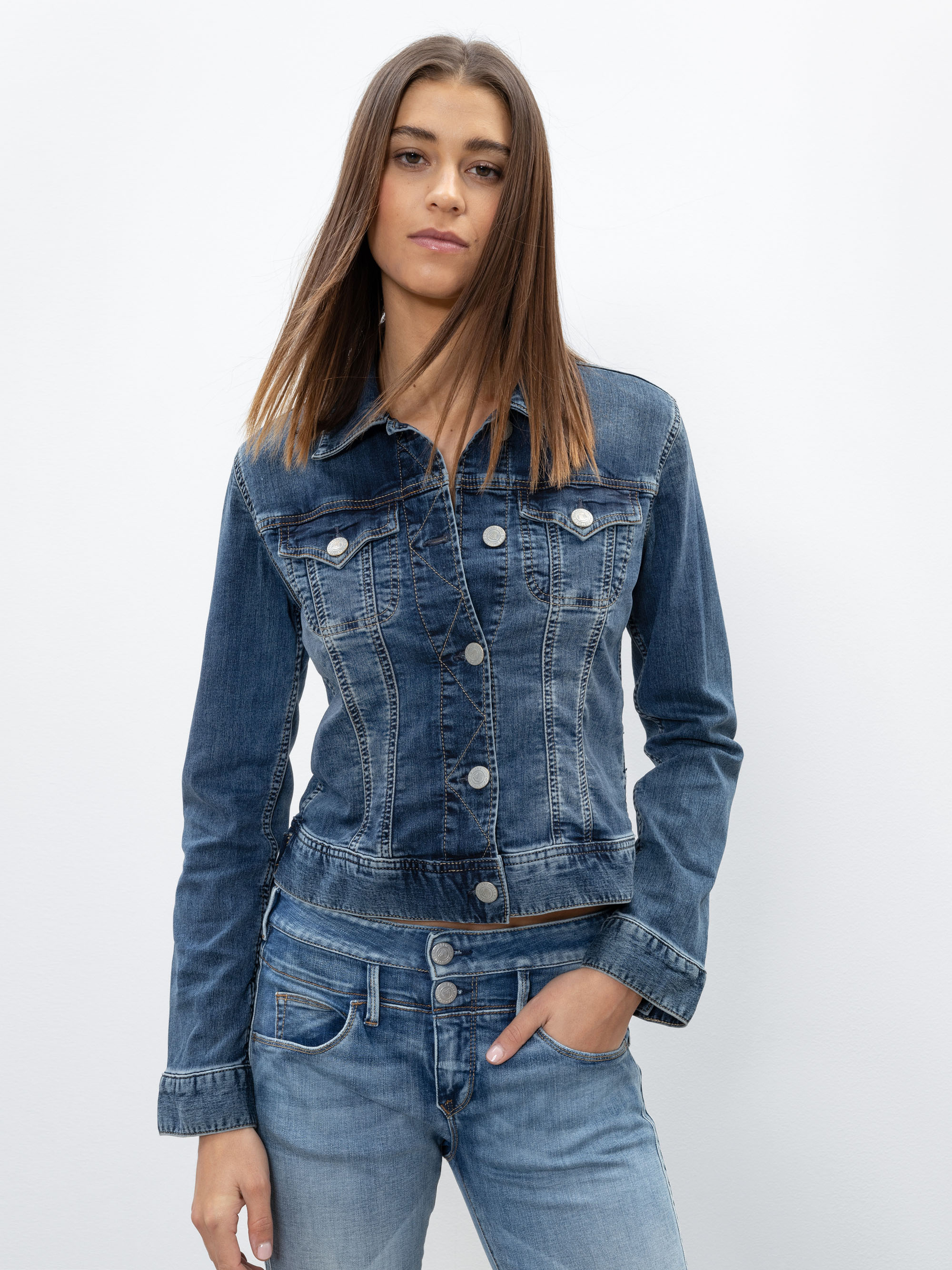 Eine Person mit glattem braunem Haar trägt eine taillierte blaue Jeansjacke und hellblaue Jeans, steht vor einem schlichten weißen Hintergrund und hat eine Hand in der Tasche.