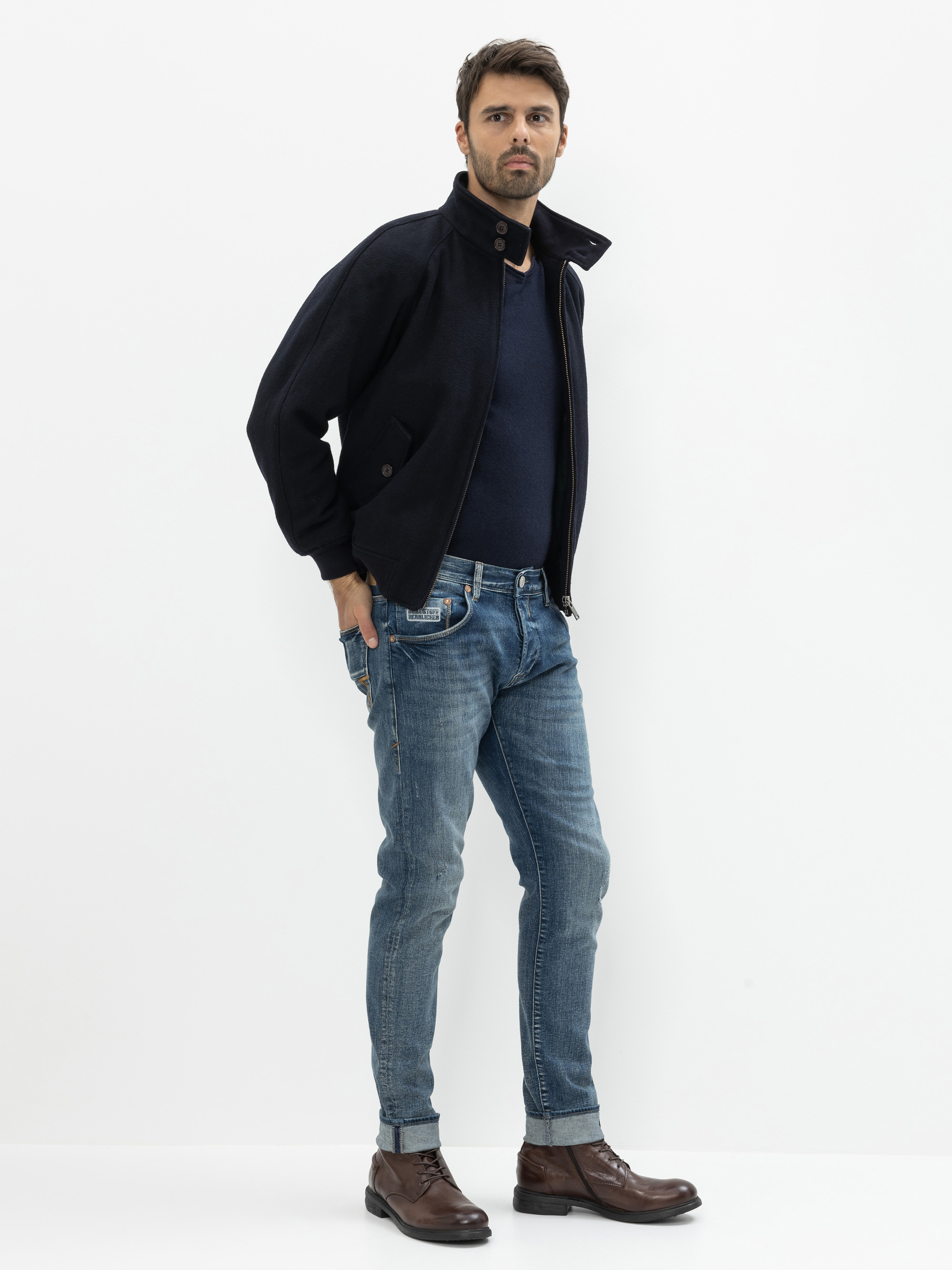 Ein Mann steht vor einem weißen Hintergrund und trägt eine dunkle Jacke über einem marineblauen Hemd, blaue Jeans und braune Lederstiefel. Er blickt nach vorne, eine Hand in der Hosentasche, die andere an der Seite liegend.