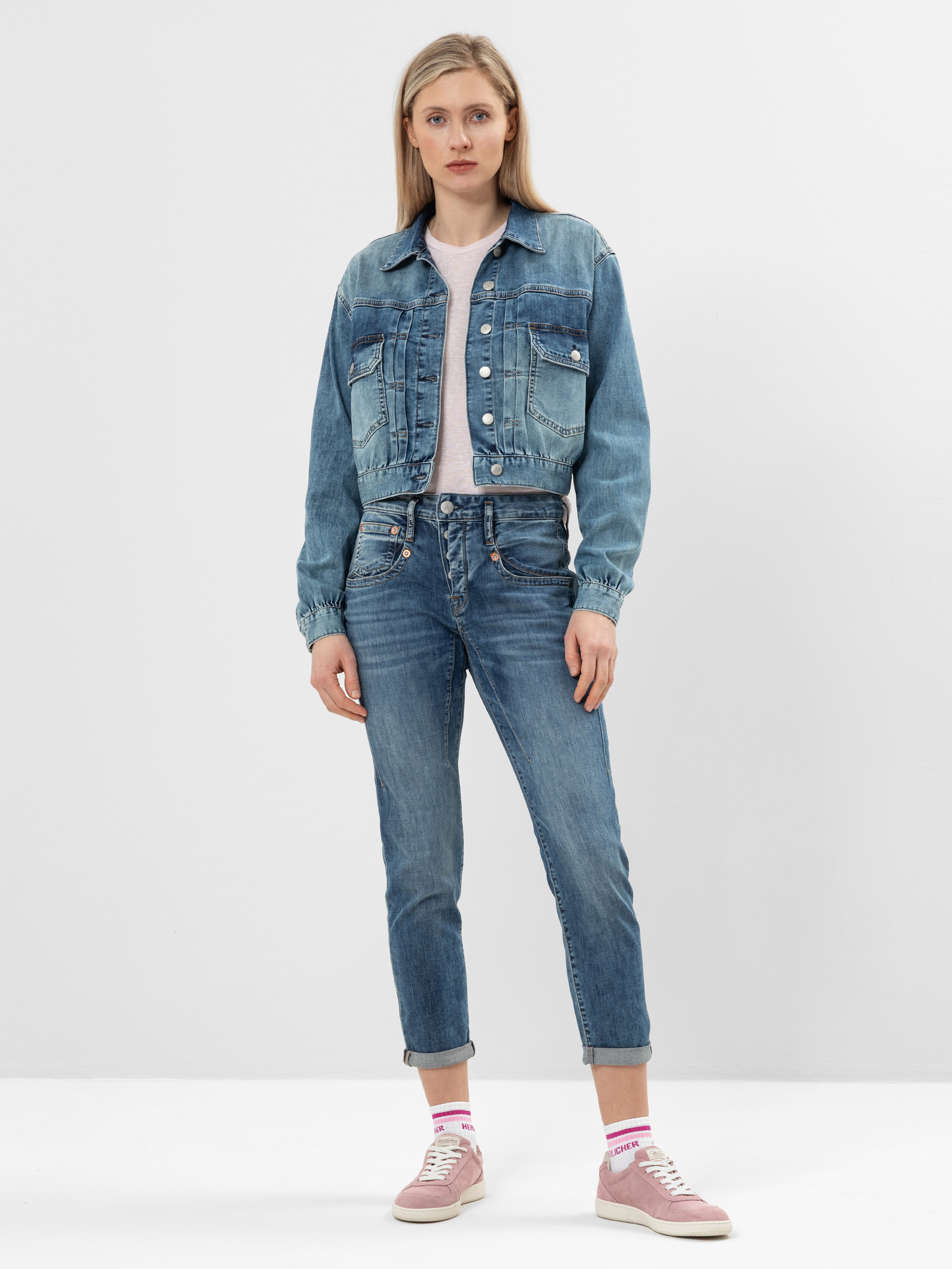 Eine Frau steht vor einem schlichten weißen Hintergrund und trägt ein hellrosa T-Shirt, eine blaue Jeansjacke, blaue Jeans, rosa Socken und rosa Turnschuhe. Sie hat glattes blondes Haar und einen neutralen Gesichtsausdruck.