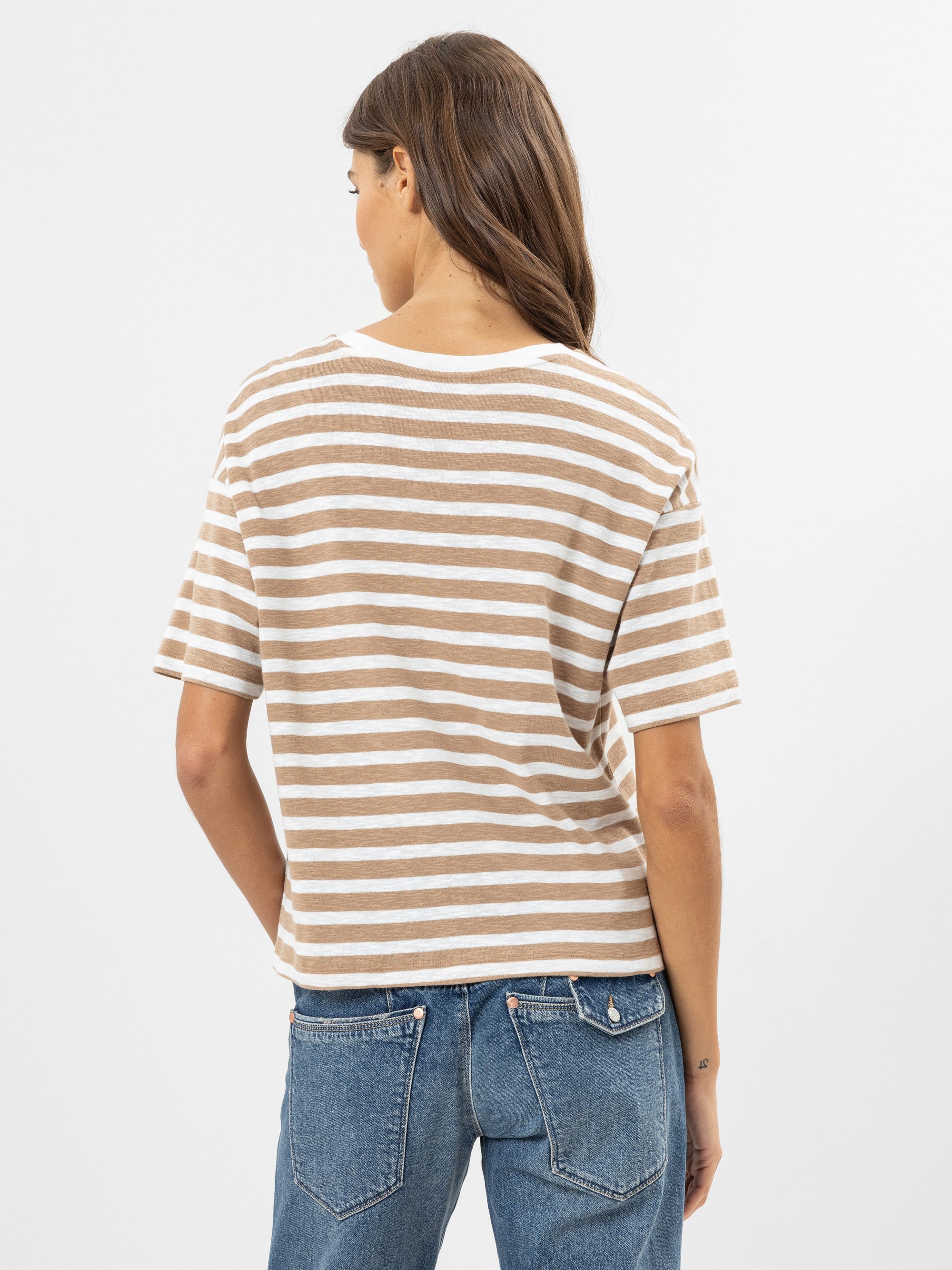 Eine Frau mit langen braunen Haaren steht mit dem Rücken zur Kamera, trägt ein braun-weiß gestreiftes kurzärmeliges Hemd und blaue Jeans vor einem schlichten weißen Hintergrund.